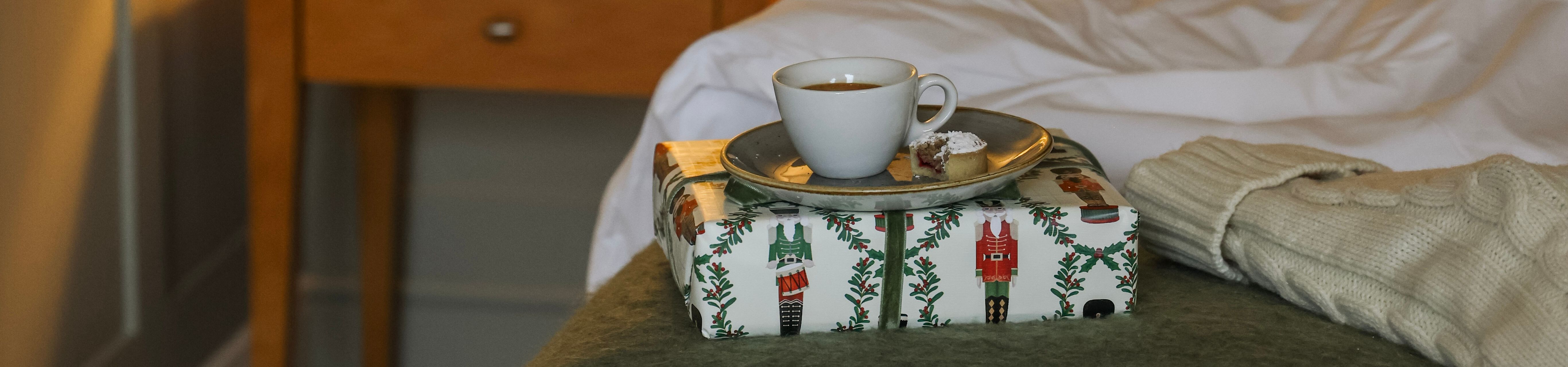 Cozy bedroom scene with a bedside lamp, a cup of coffee on a wrapped gift, and a bed with white pillows and a green throw.