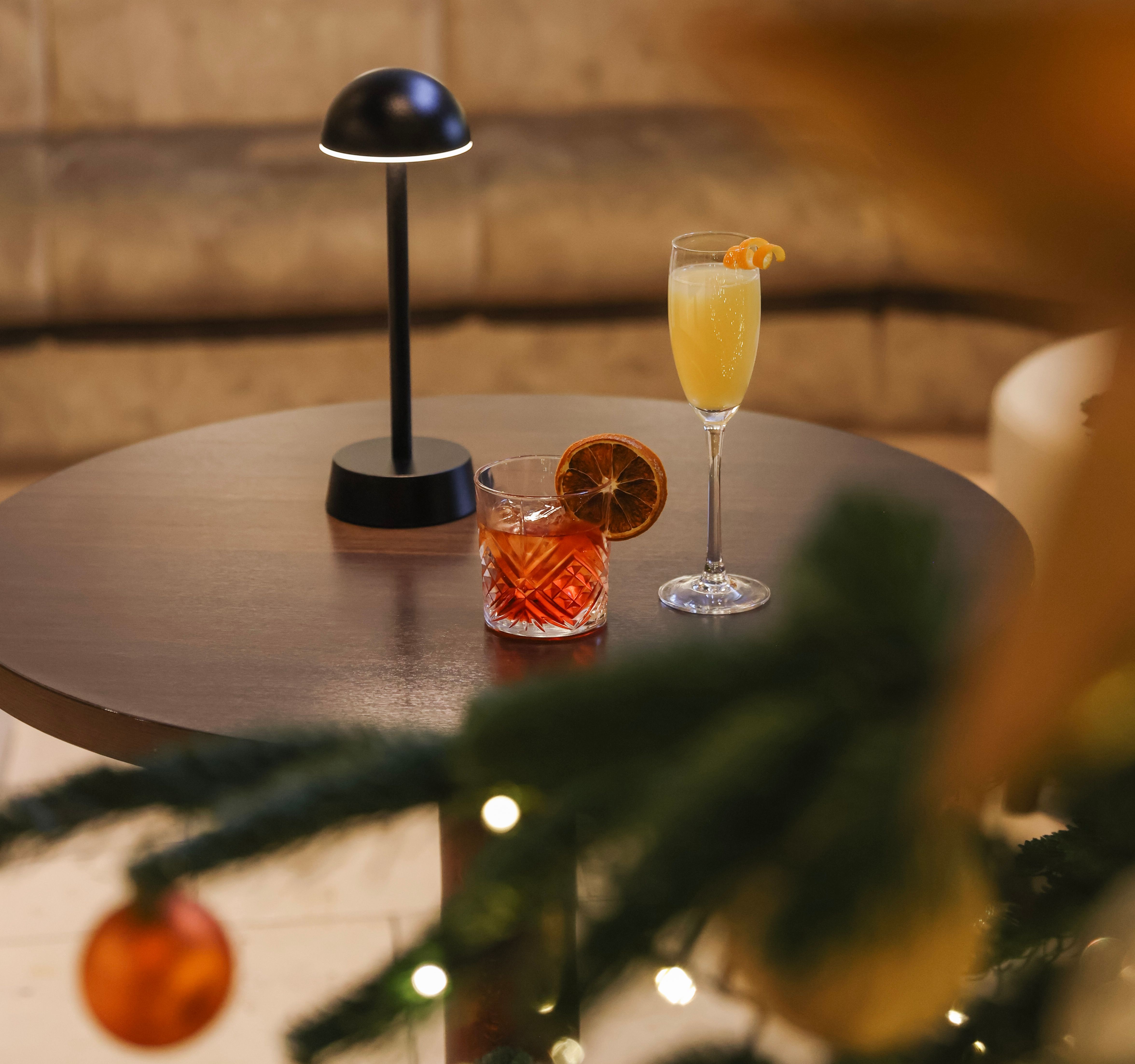 Round table with two cocktails beside a small lamp, with a decorated Christmas tree and festive ornaments in the foreground.