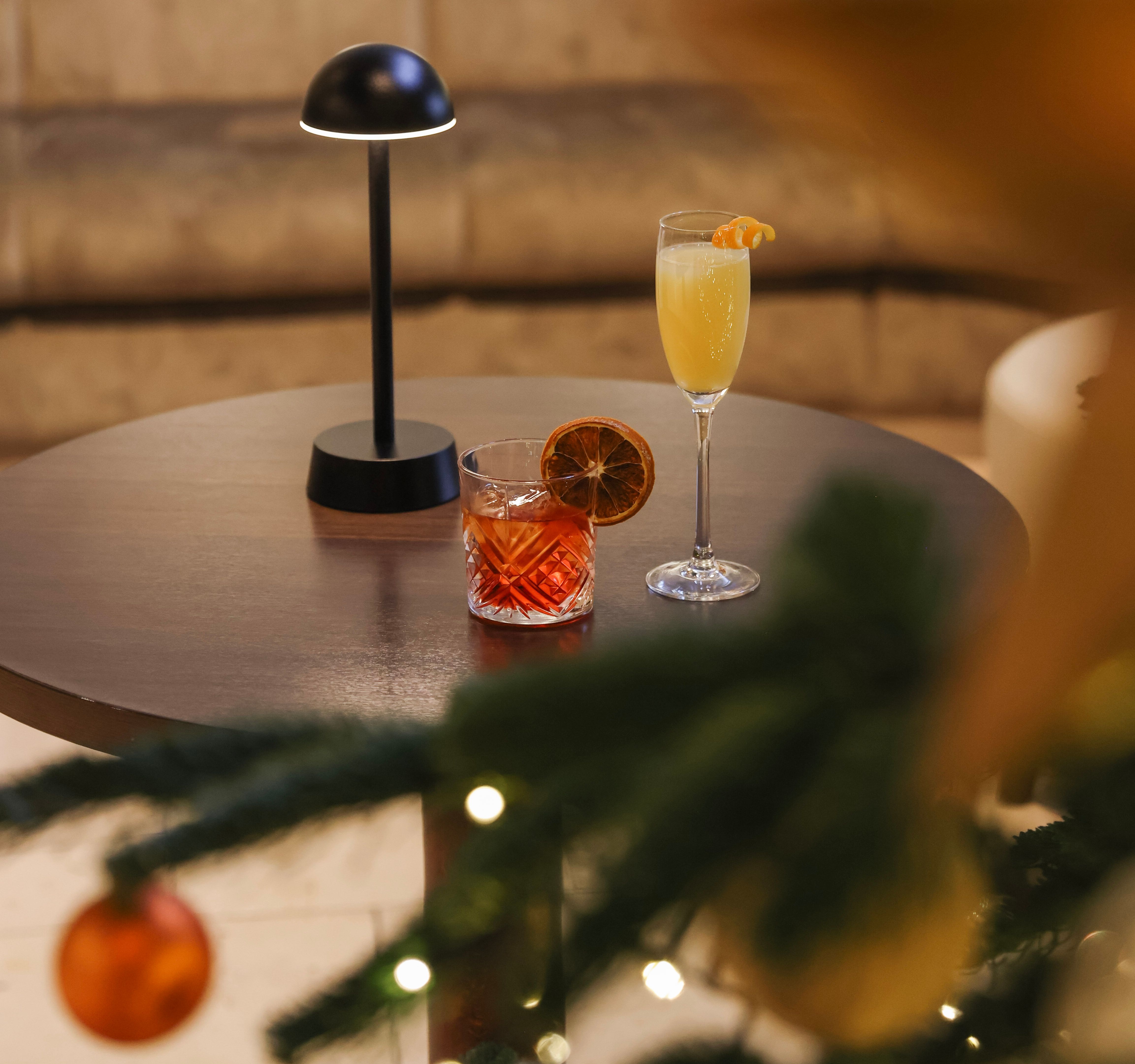 Round table with two cocktails beside a small lamp, with a decorated Christmas tree and festive ornaments in the foreground.