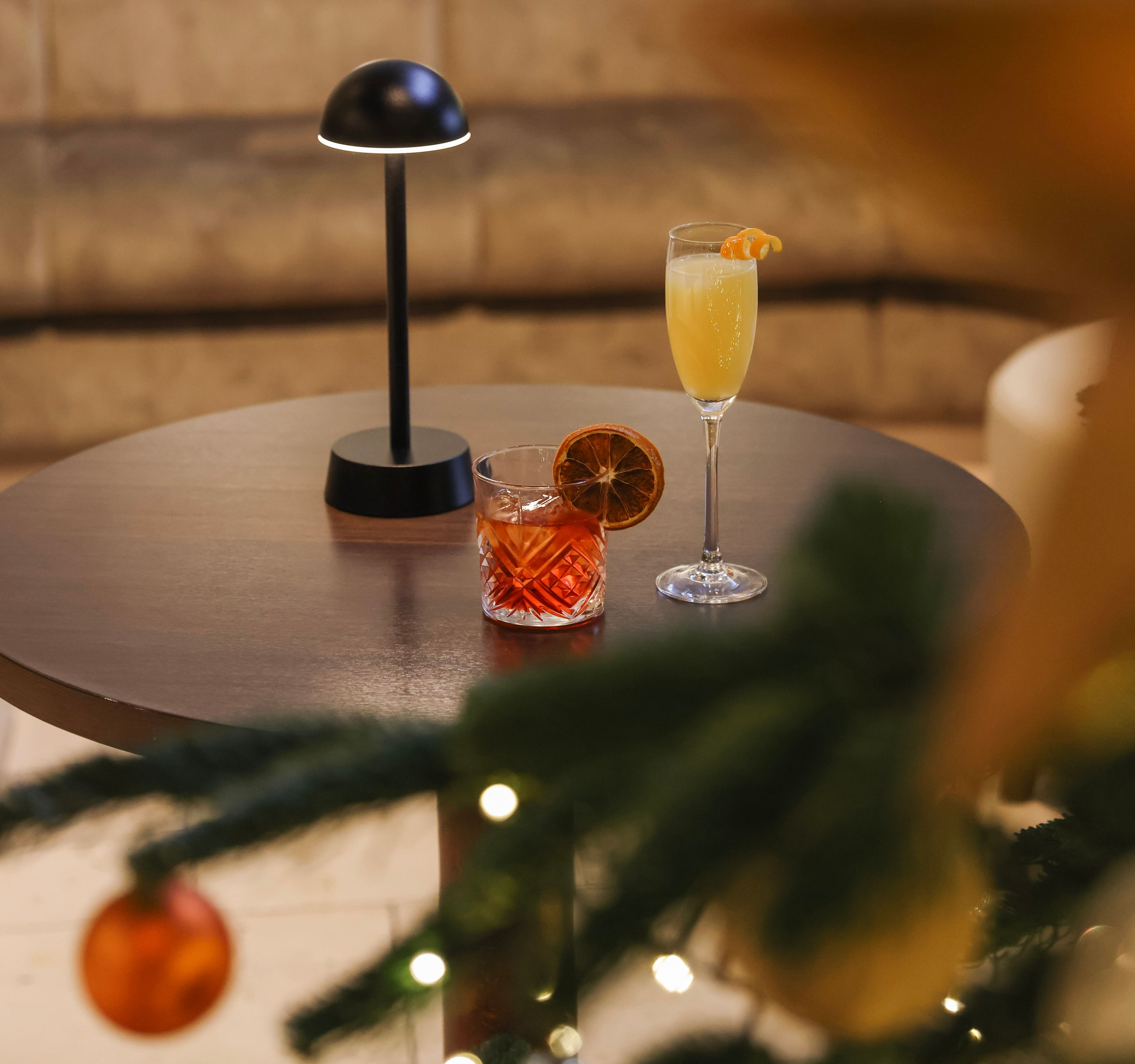 Round table with two cocktails beside a small lamp, with a decorated Christmas tree and festive ornaments in the foreground.