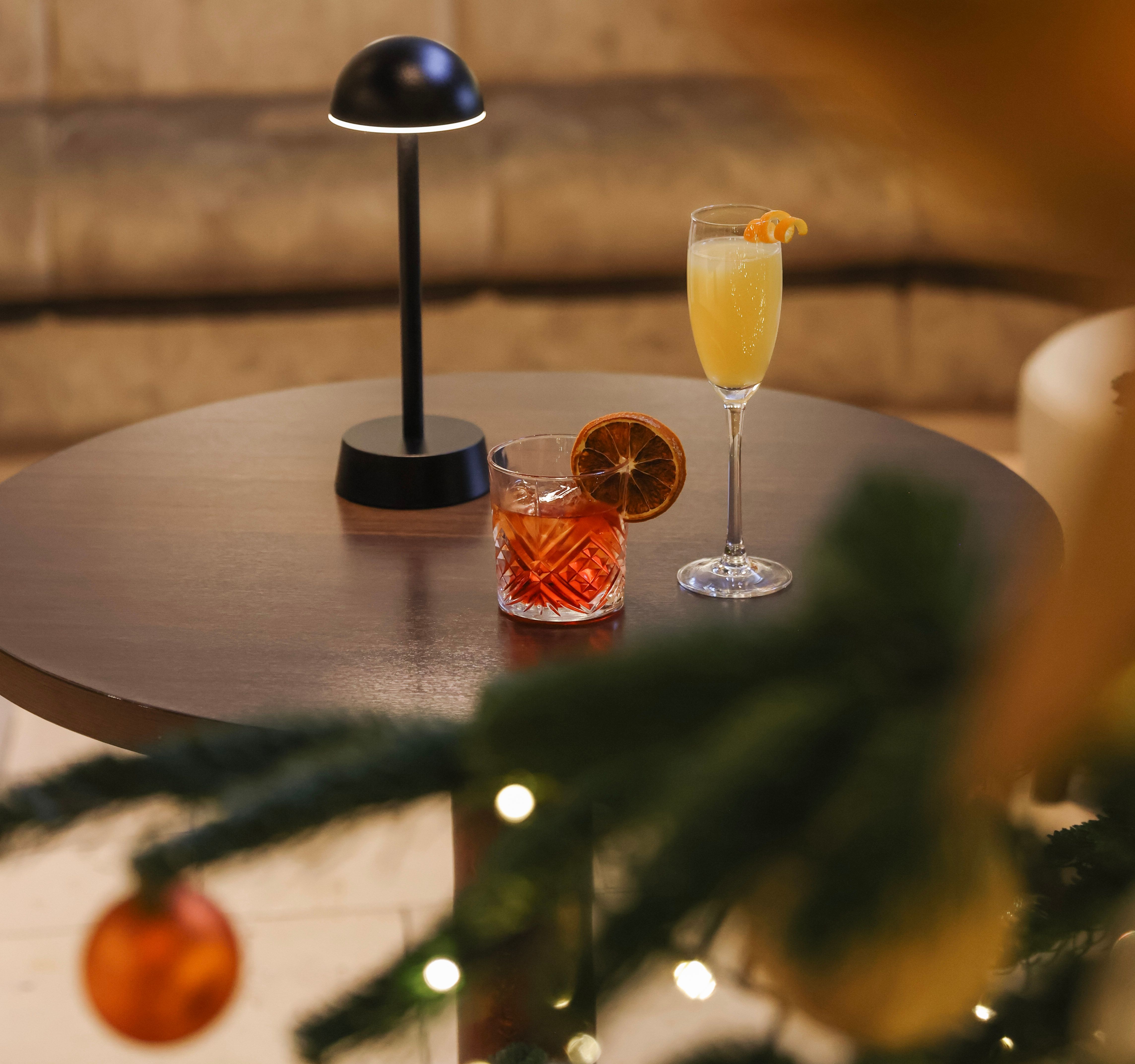 Round table with two cocktails beside a small lamp, with a decorated Christmas tree and festive ornaments in the foreground.