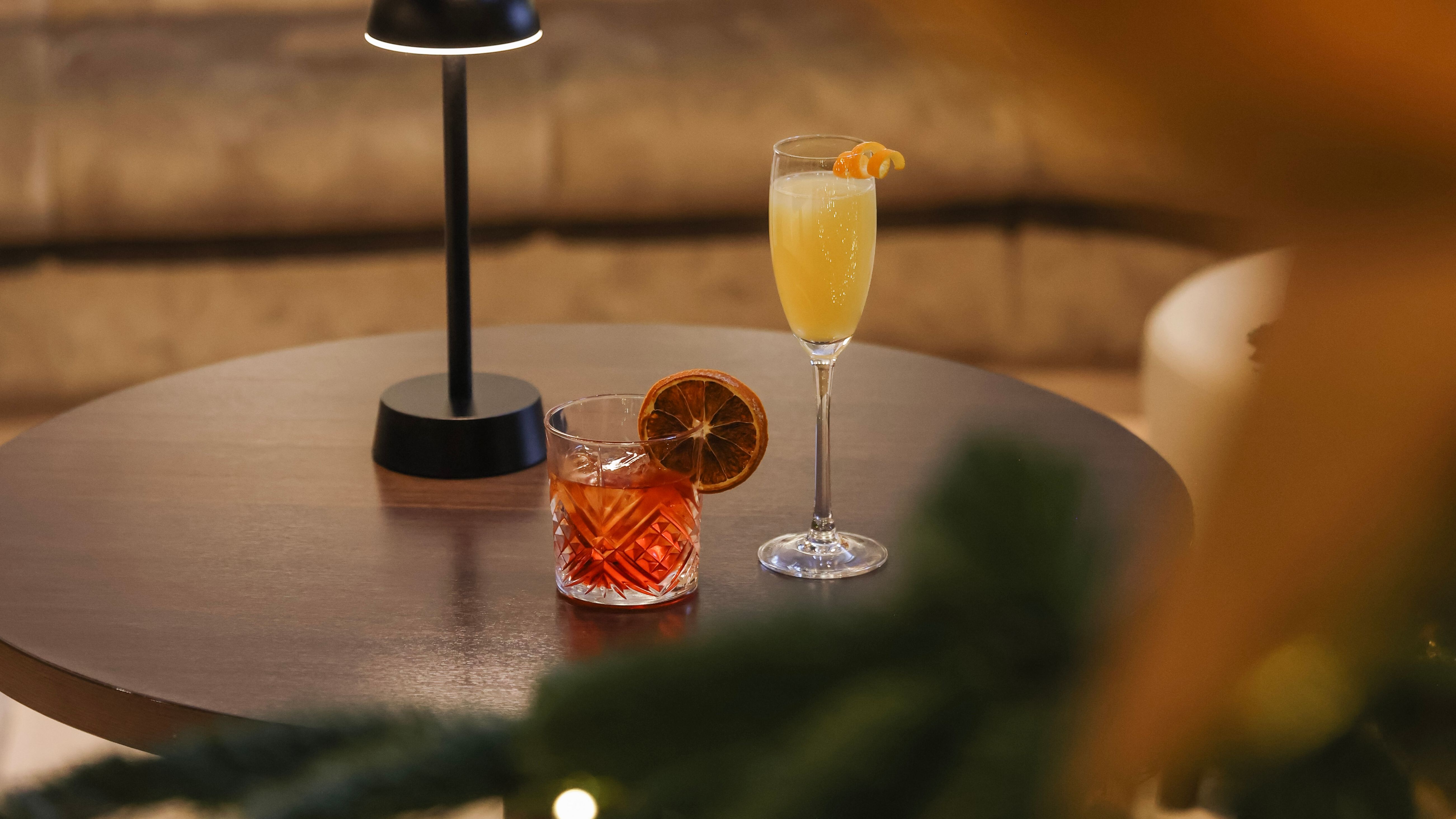 Round table with two cocktails beside a small lamp, with a decorated Christmas tree and festive ornaments in the foreground.