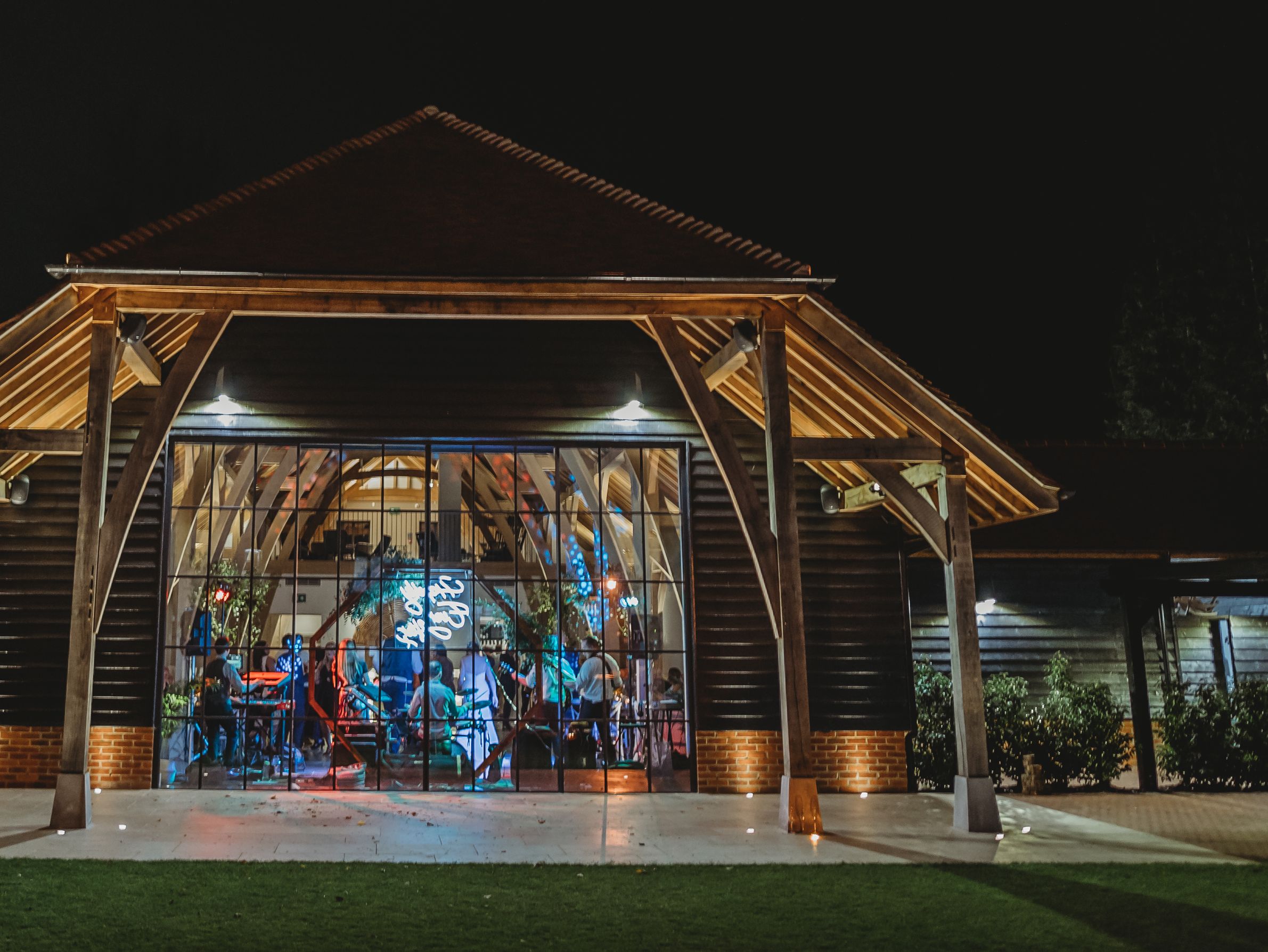 Night view of a wooden barn-like venue with glass front, warmly lit from inside, with people visible through the windows.