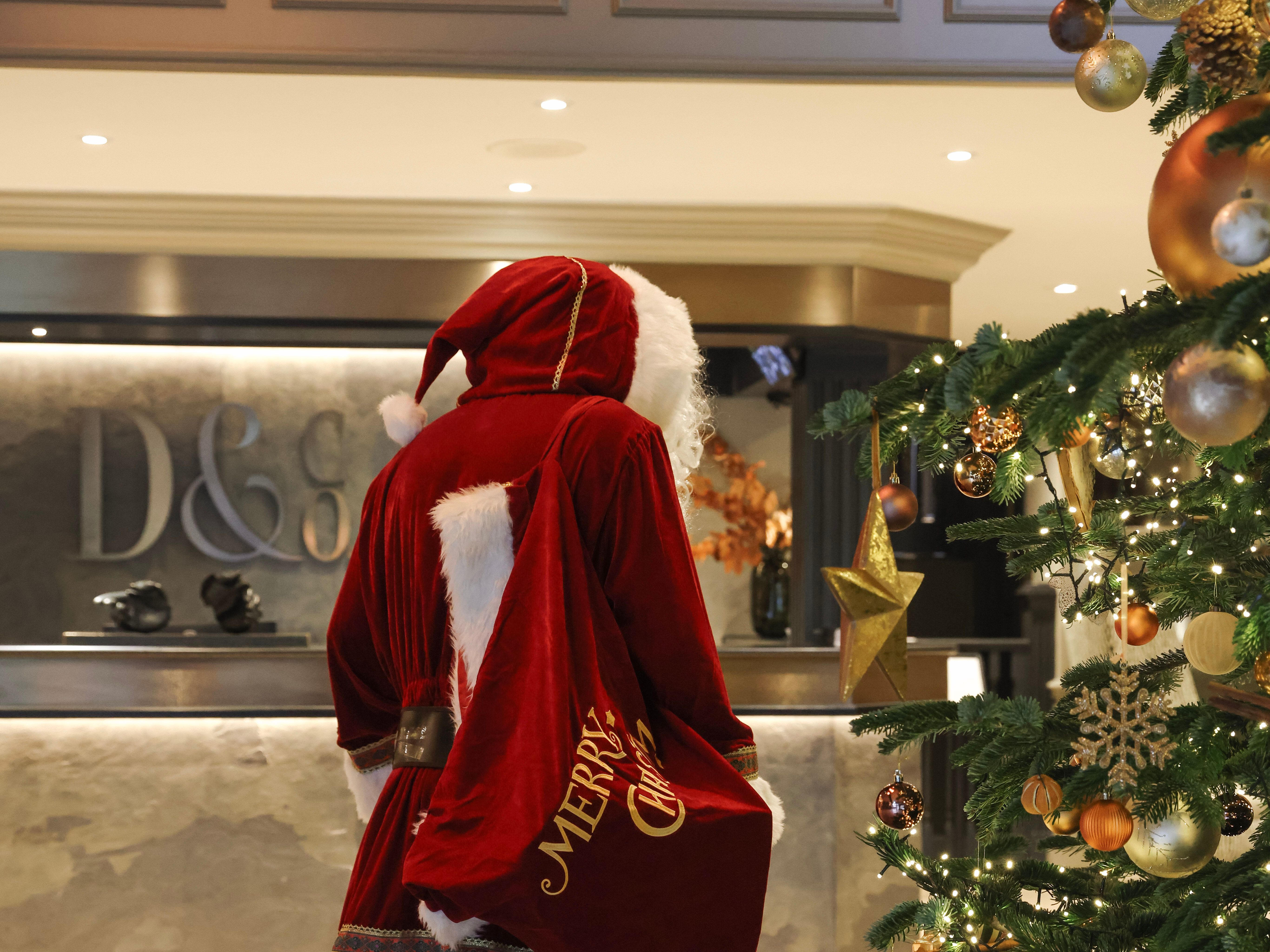 Santa Claus standing indoors next to a decorated Christmas tree with ornaments and lights.