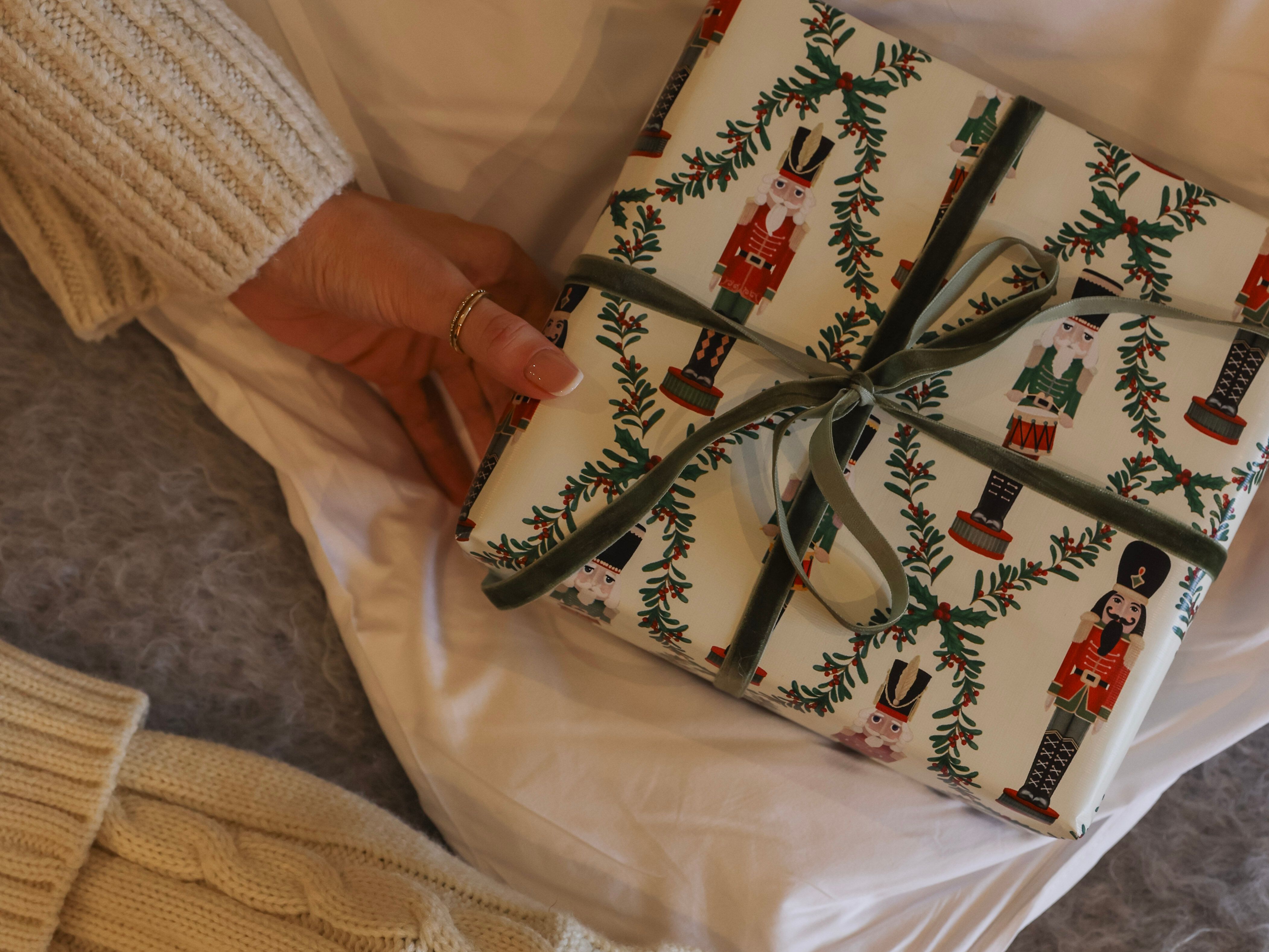 Hand holding a wrapped Christmas present next to a cream-colored knitted stocking on a cozy surface