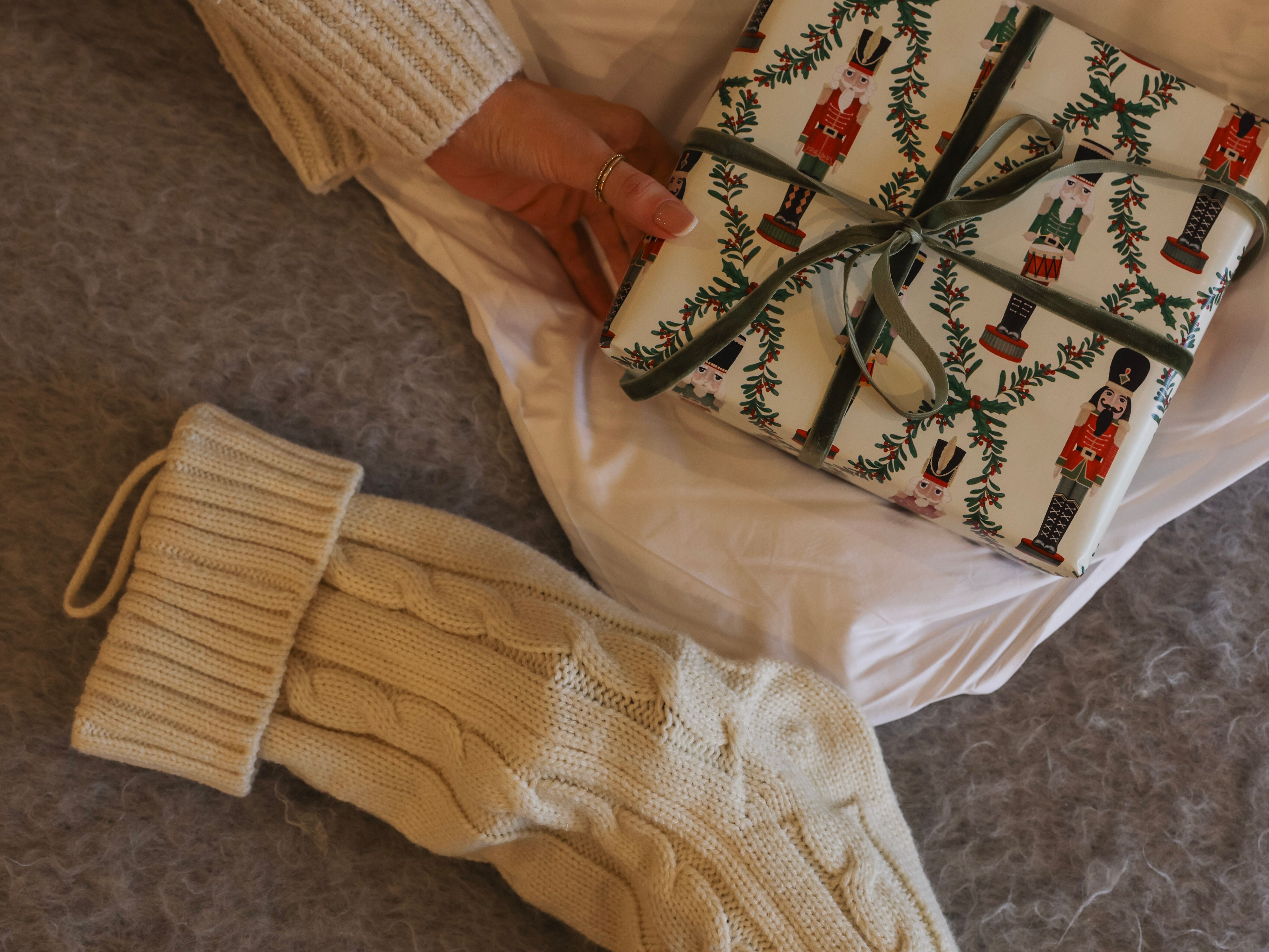 Hand holding a wrapped Christmas present next to a cream-colored knitted stocking on a cozy surface