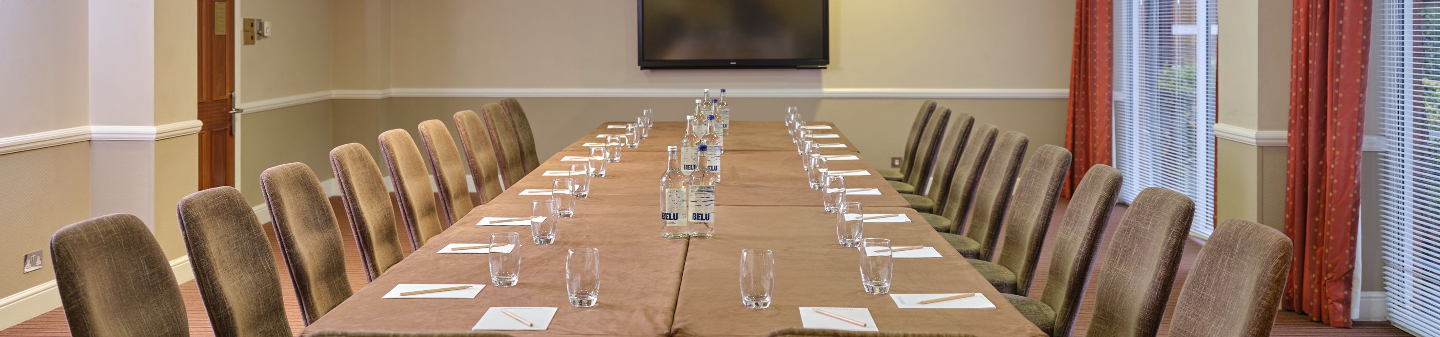 Conference room set up with U-shaped table arrangement, chairs, water bottles, glasses, notepads, and a television screen on the wall.