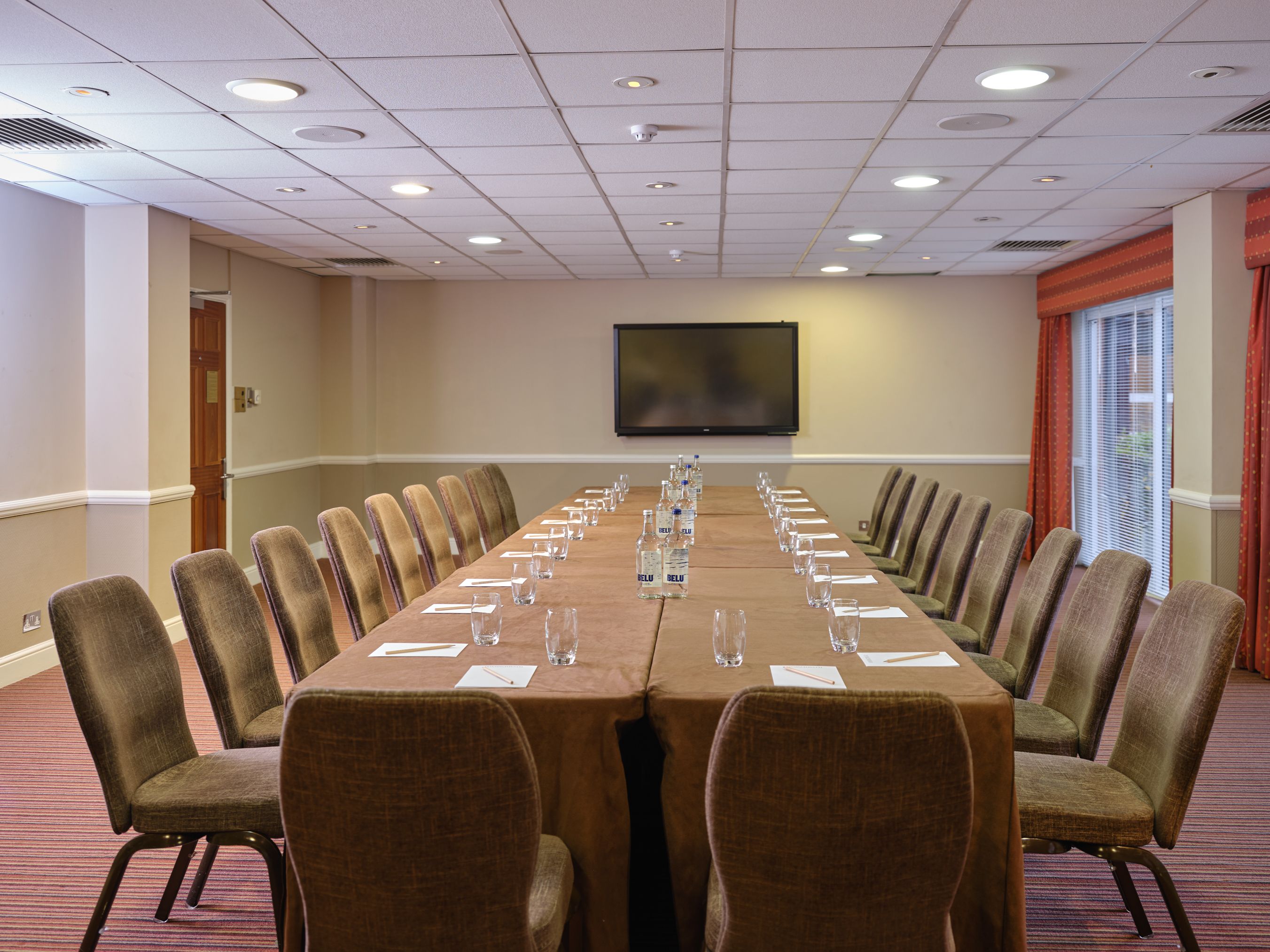 Conference room set up with U-shaped table arrangement, chairs, water bottles, glasses, notepads, and a television screen on the wall.