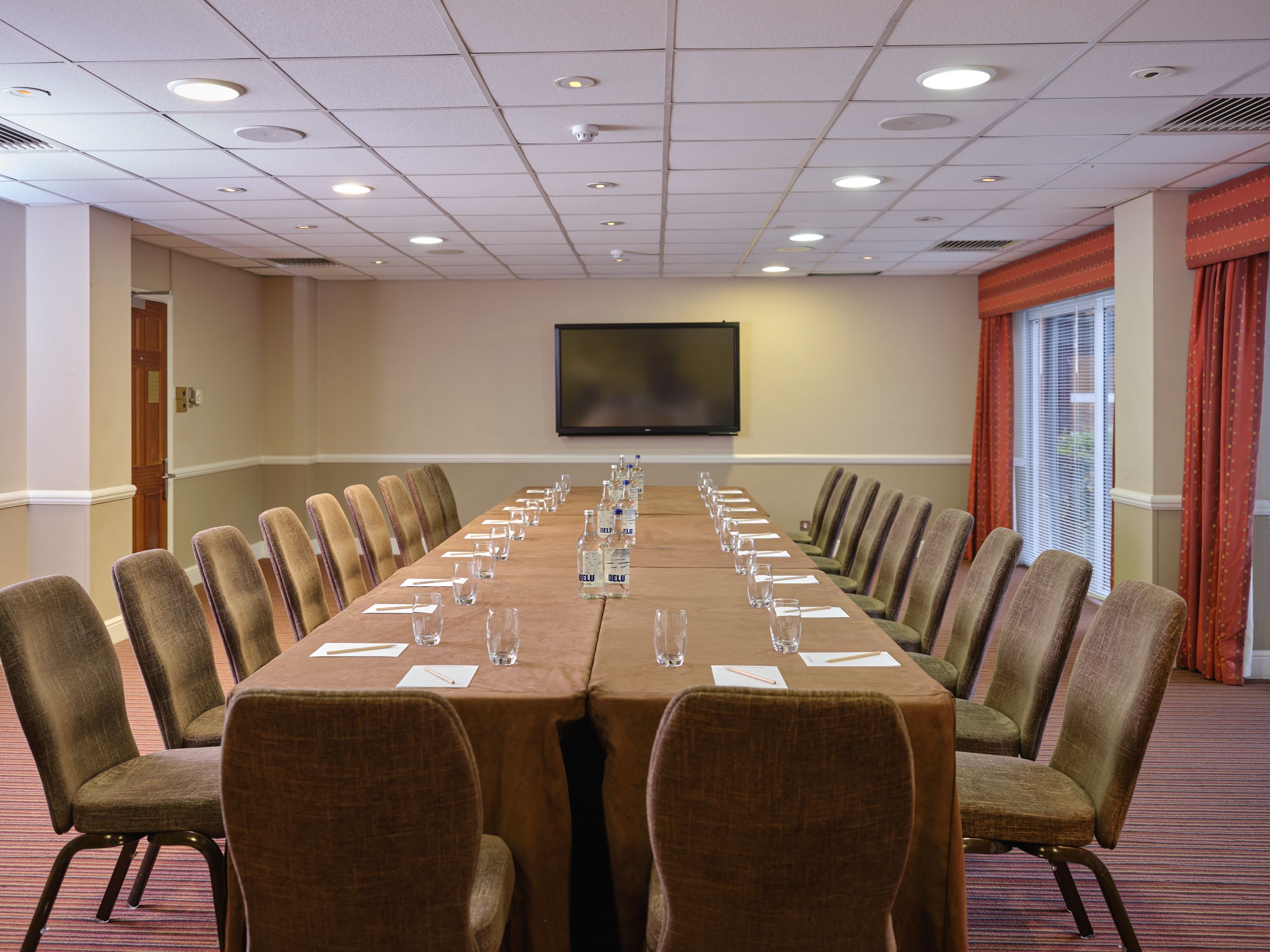 Boardroom style conference room with a long table, brown chairs, water bottles, glasses, notepads, and a wall-mounted TV screen.