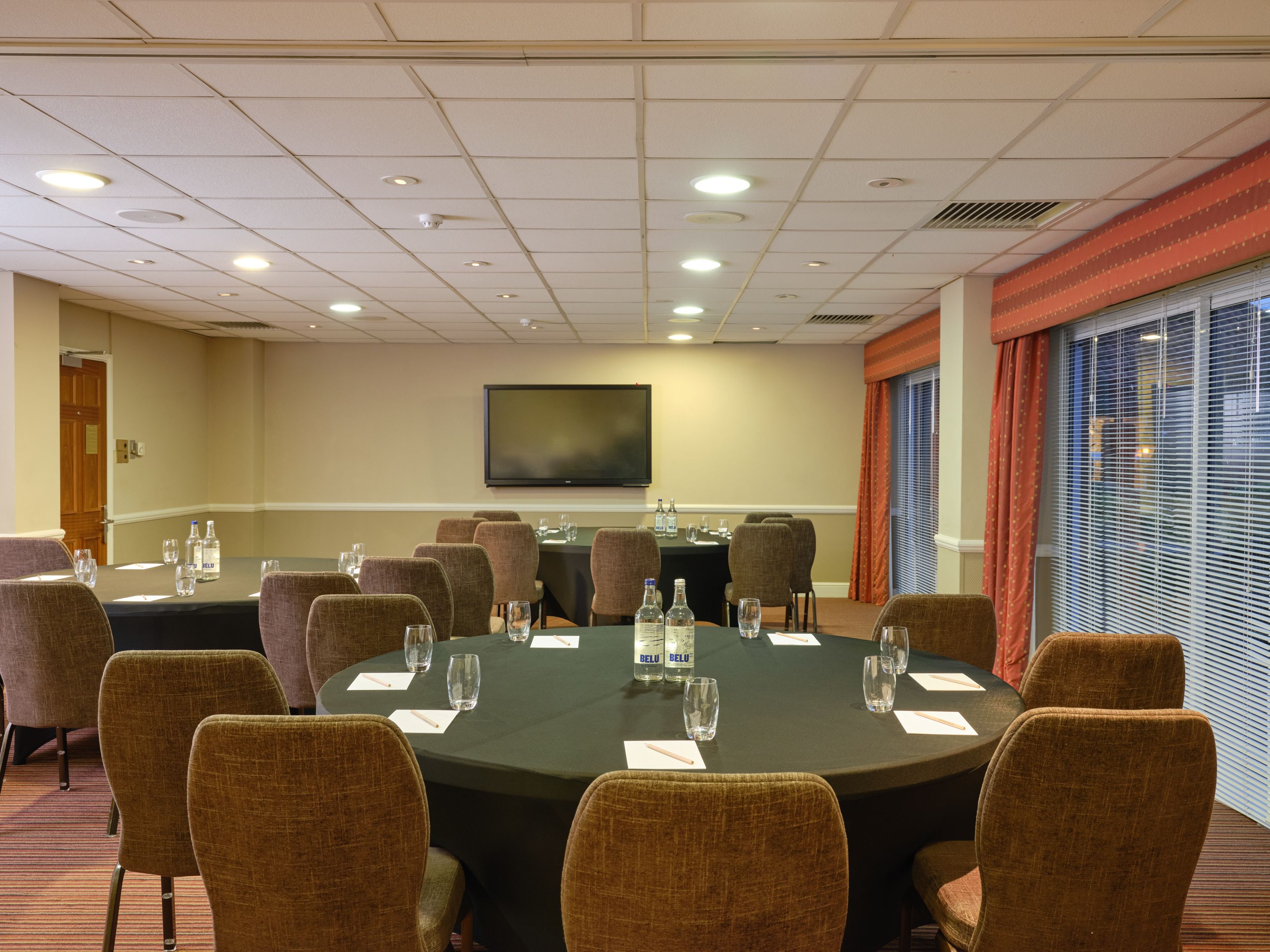 Conference room with round tables, brown chairs, water bottles, glasses, notepads, and a large wall-mounted TV.