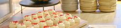 Assorted mini desserts on a wooden tray with plates and fruit bowl in the background