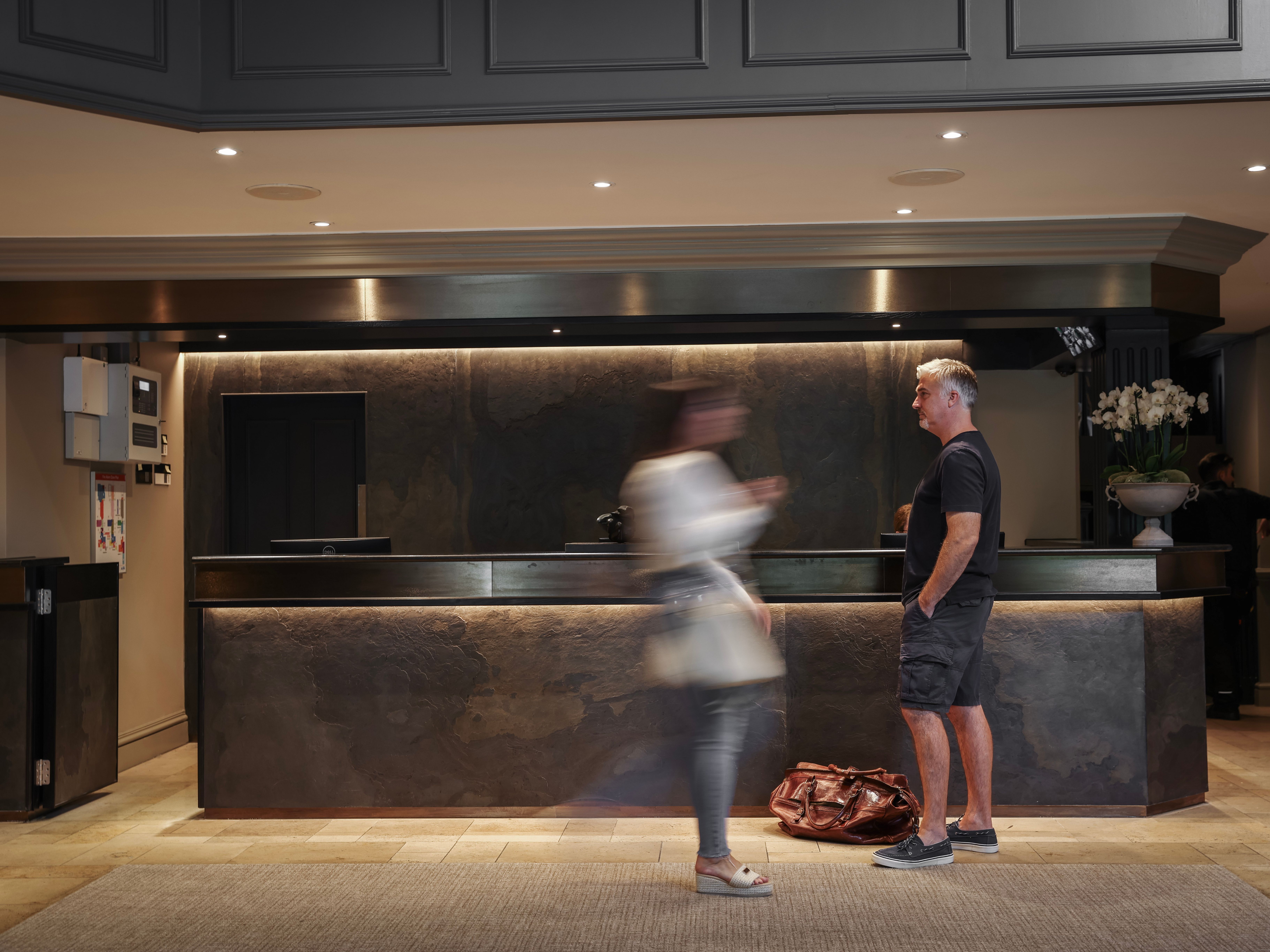 A modern hotel reception area with a man standing near the counter and a blurred woman walking by.