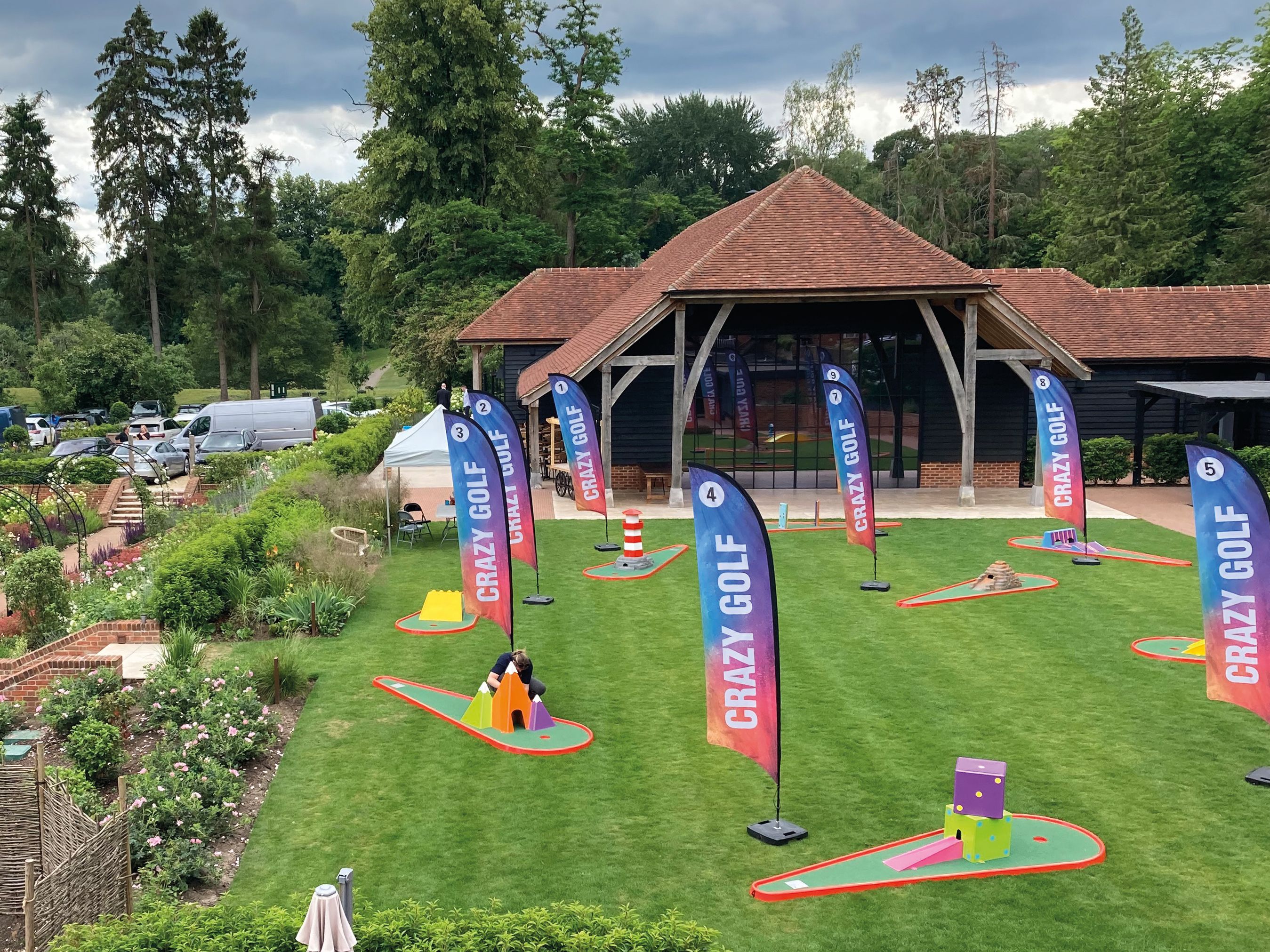 Outdoor mini golf course with colorful obstacles and CRAZY GOLF banners, garden paths, and a building in the background.