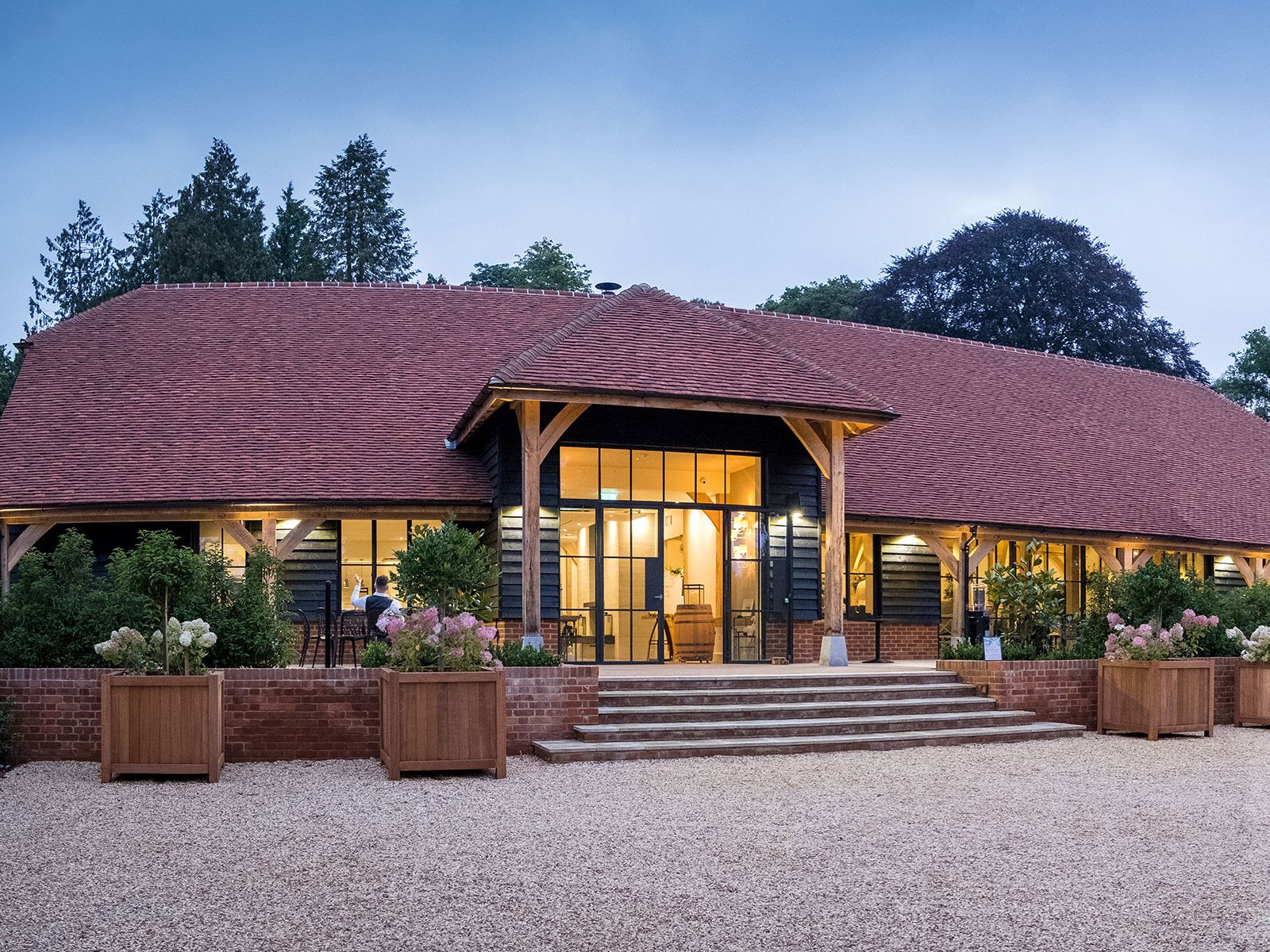A large, rustic building with a red-tiled roof, large wooden beams, and bright interior lighting, surrounded by greenery at dusk.