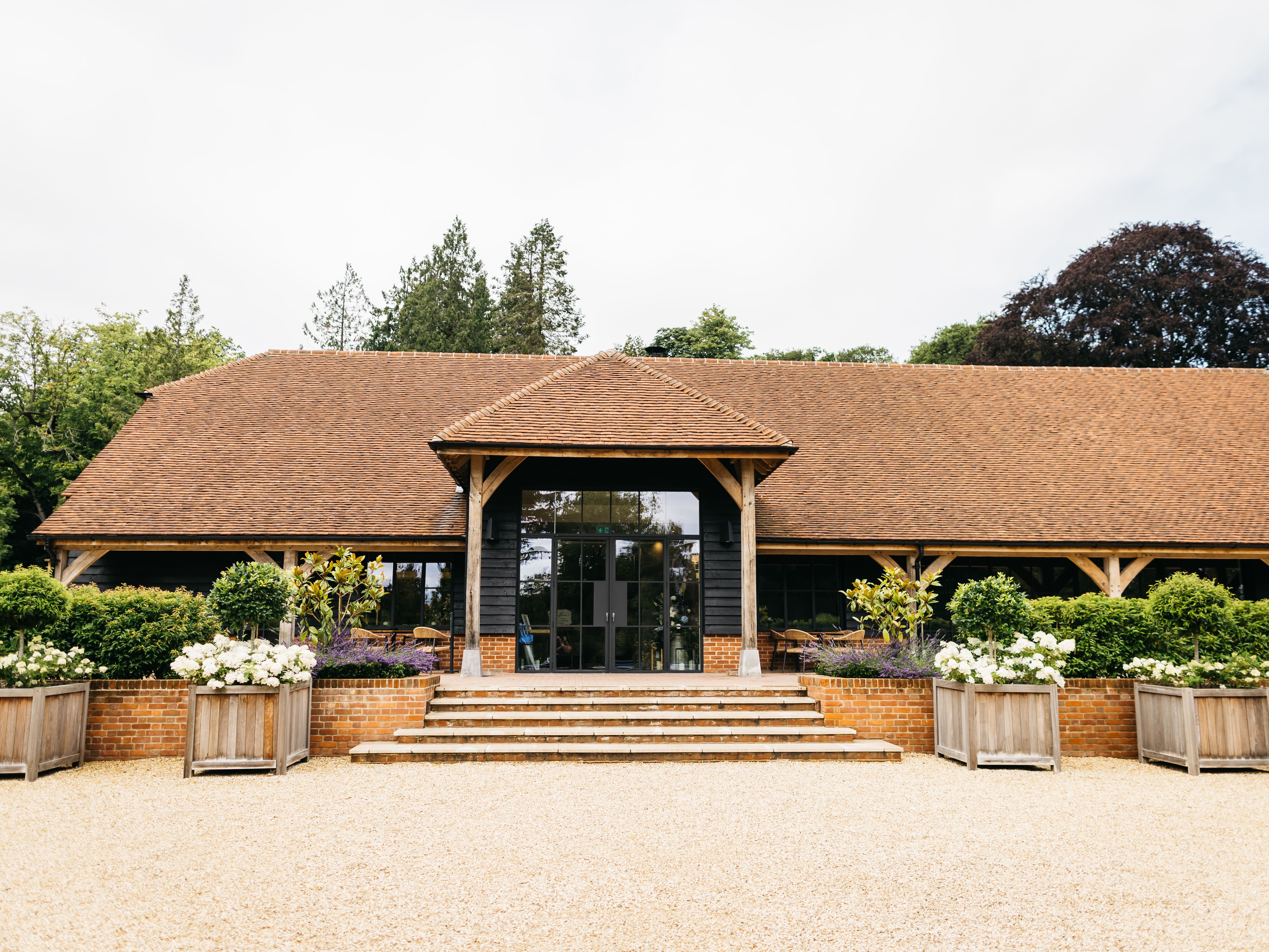 A rustic barn with a tiled roof and large glass doors, surrounded by greenery and planter boxes with flowers.