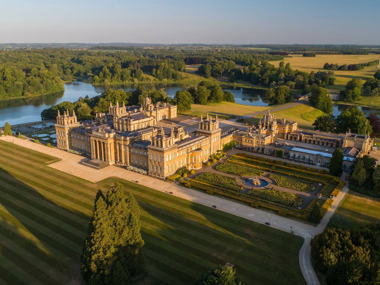 Aerial view of a grand palace with formal gardens beside a lake, surrounded by parkland