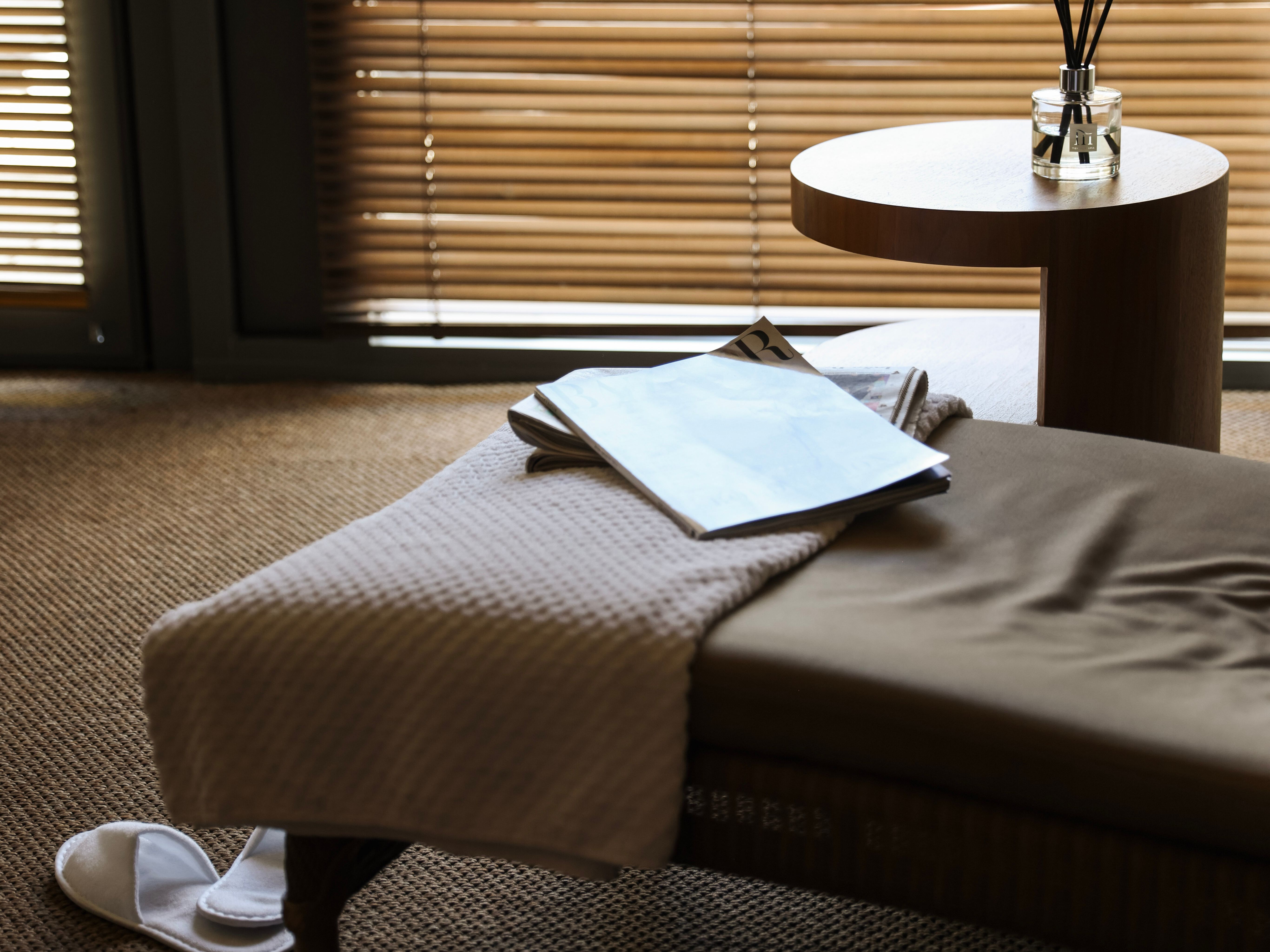Sunlit bedroom corner with a bedside table, reed diffuser and open magazine beside a bed