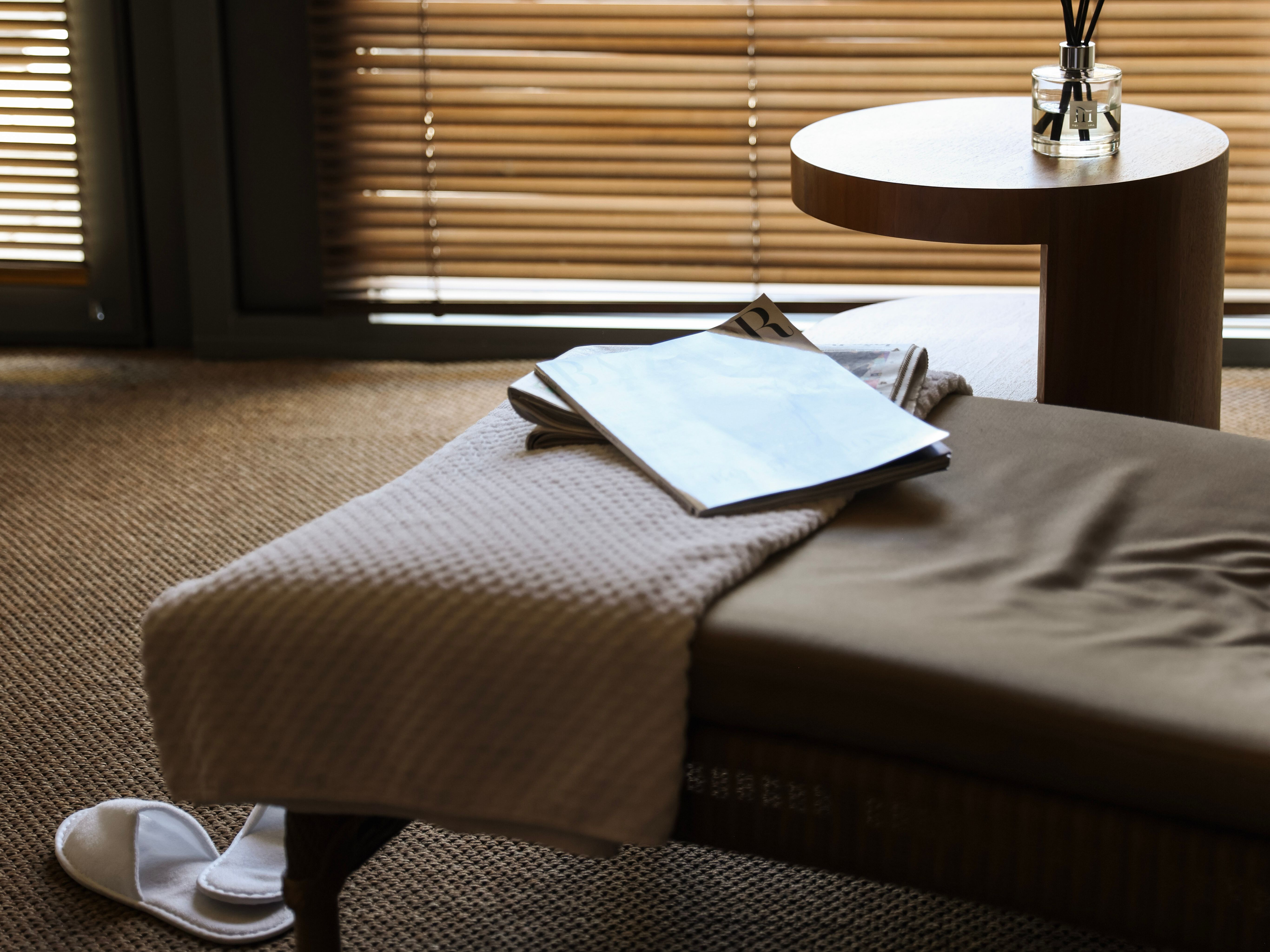 Sunlit bedroom corner with a bedside table, reed diffuser and open magazine beside a bed