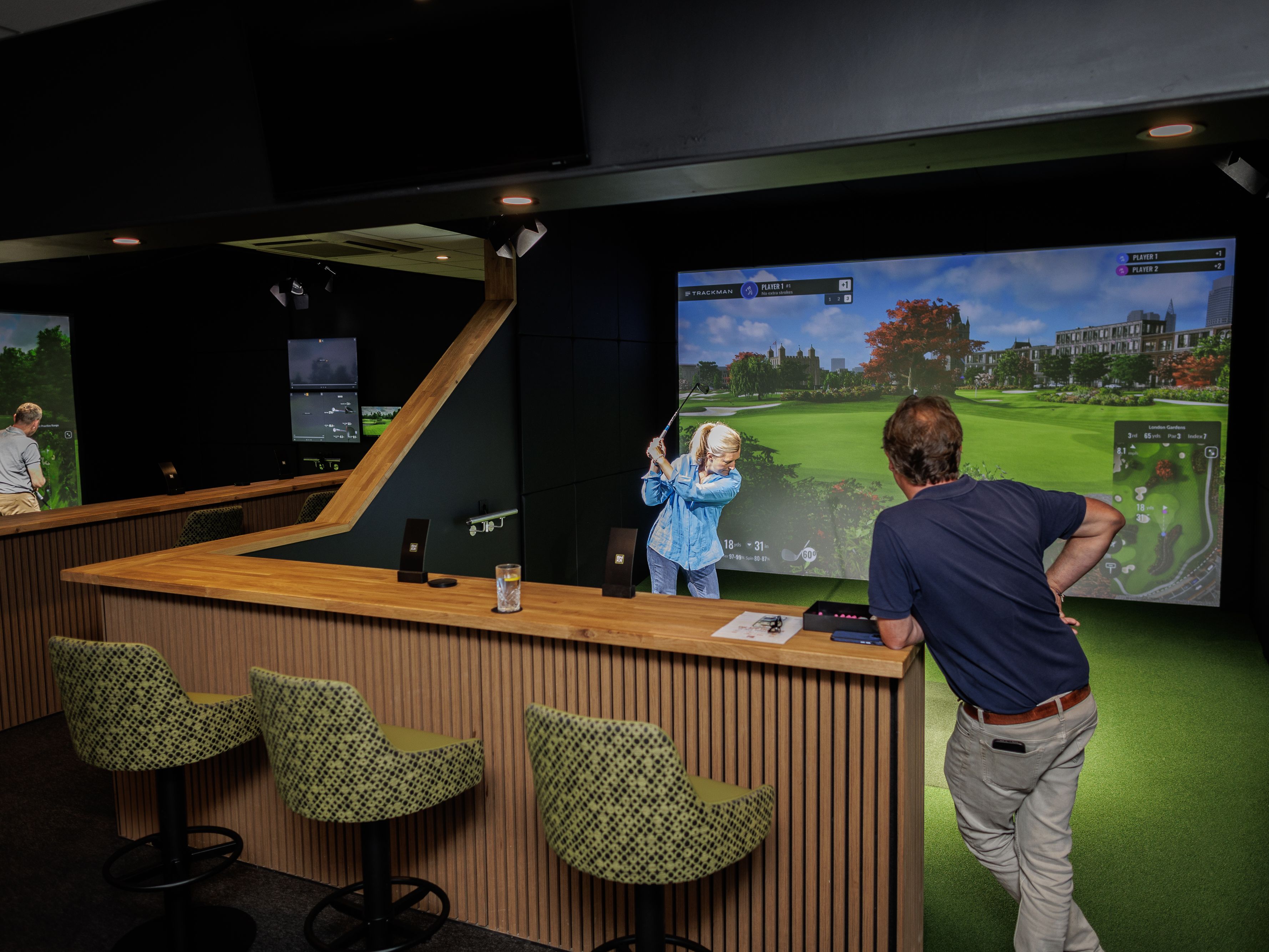 Indoor golf simulator bay with golfer swinging while another person watches from a bar counter