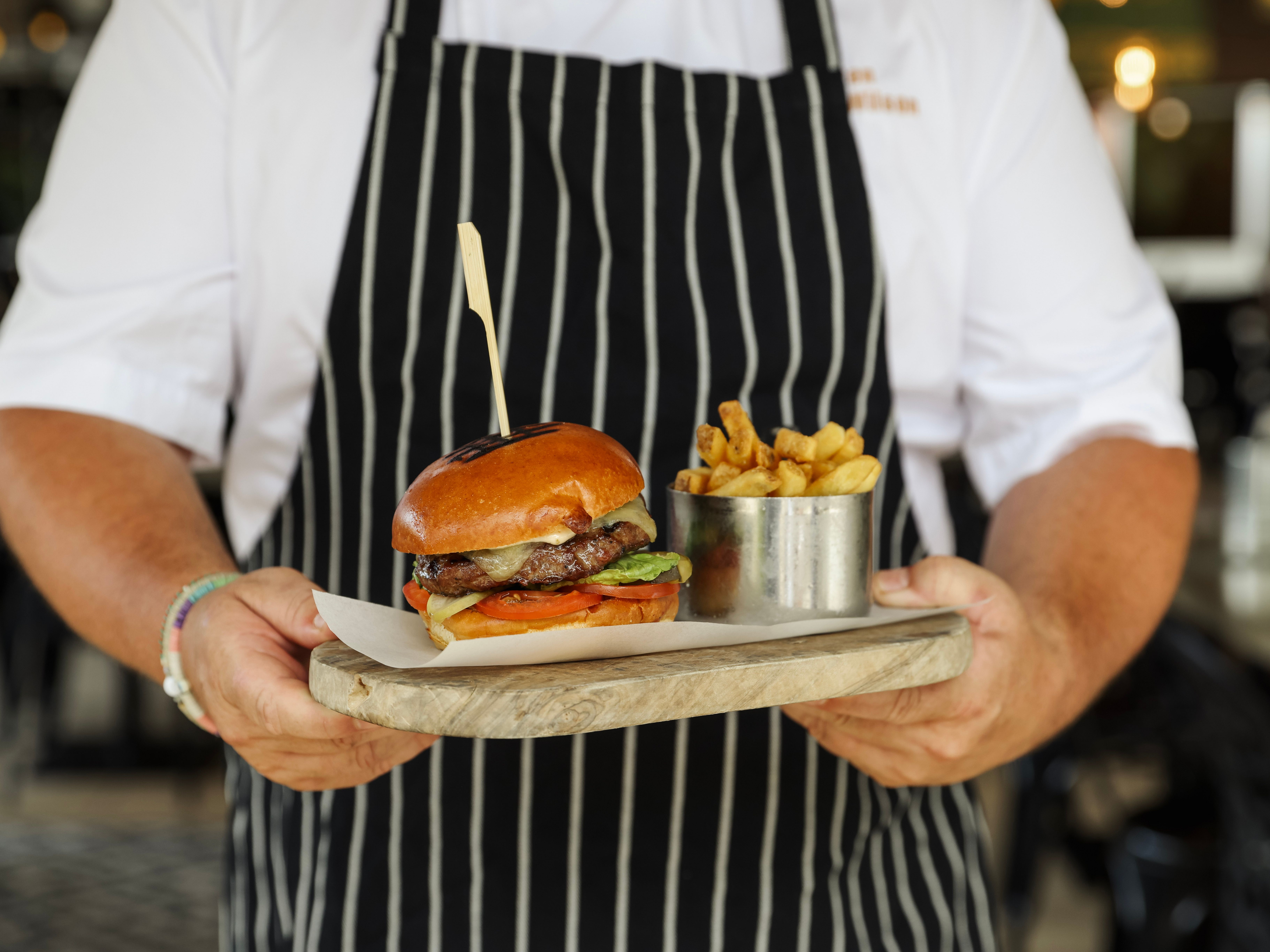 Chef holding a wooden board with a beef burger and a pot of chips
