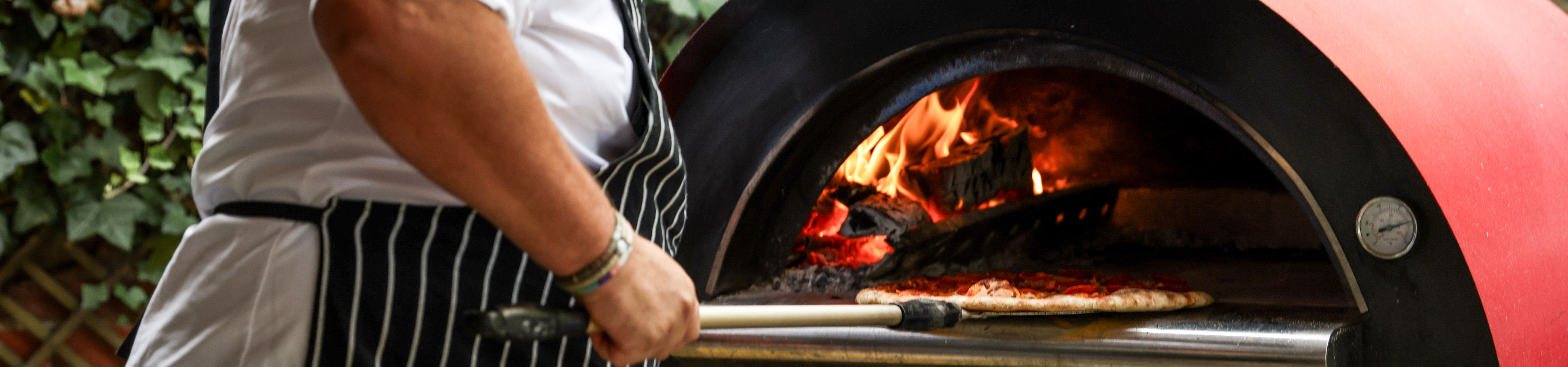 Chef in a striped apron tending pizzas in a wood-fired oven outdoors