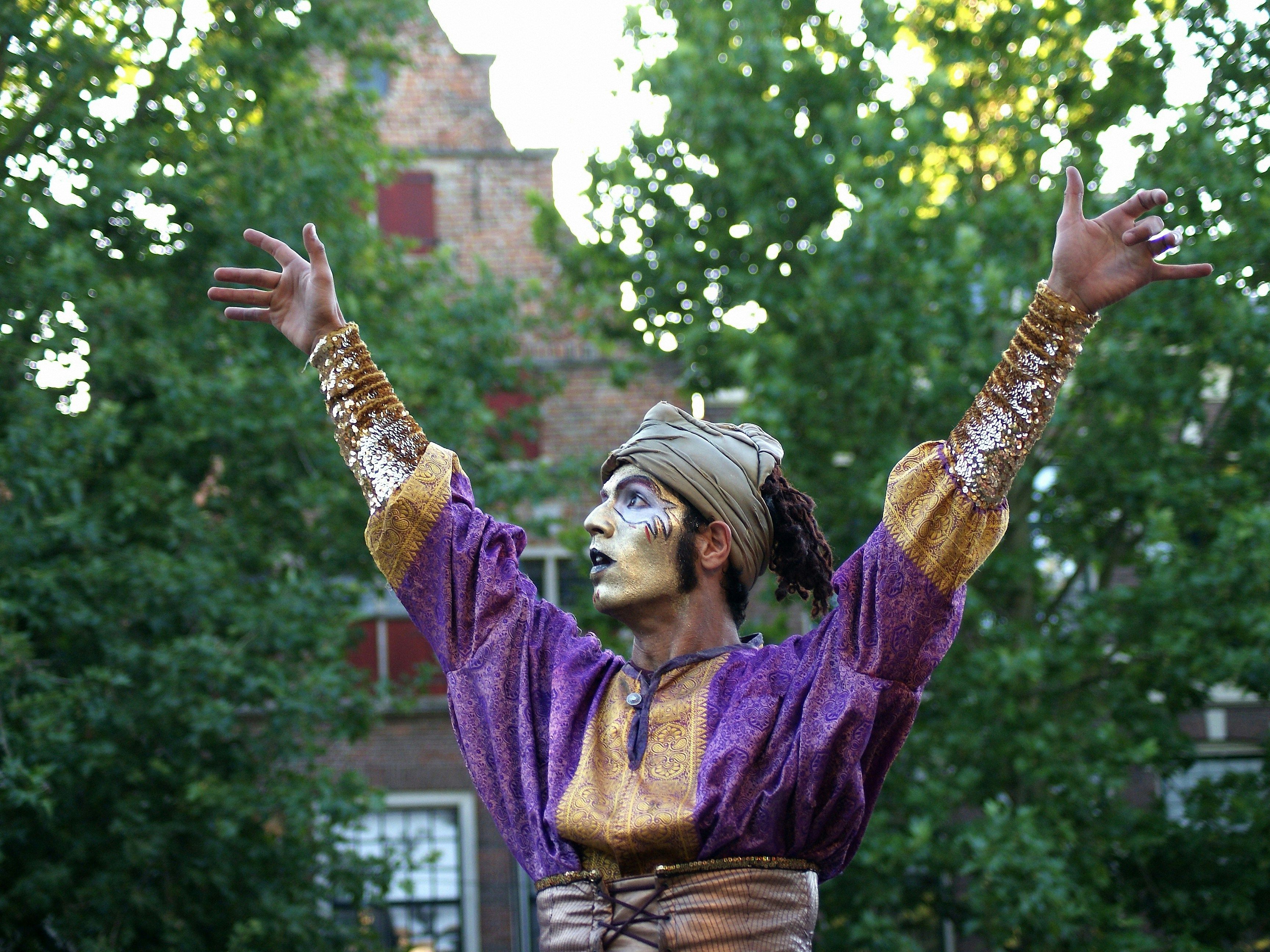 Street performer in a purple and gold costume with face paint, arms raised in a park