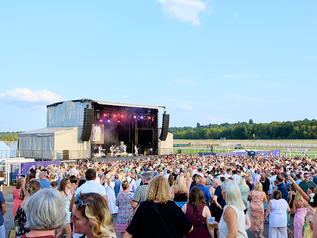 Crowd gathered in front of an outdoor stage with live band at Newbury Racecourse on a sunny summer evening