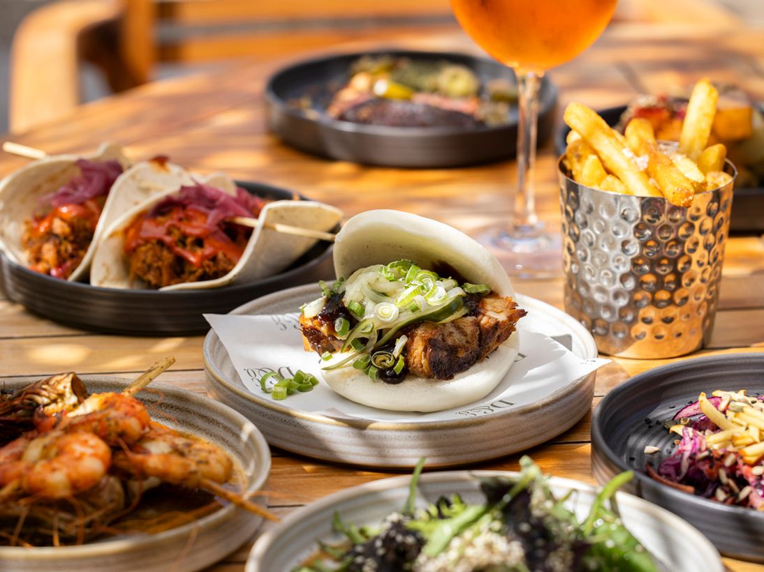 Assorted restaurant dishes on a wooden table, including a steamed bao bun, tacos, chips and a cocktail