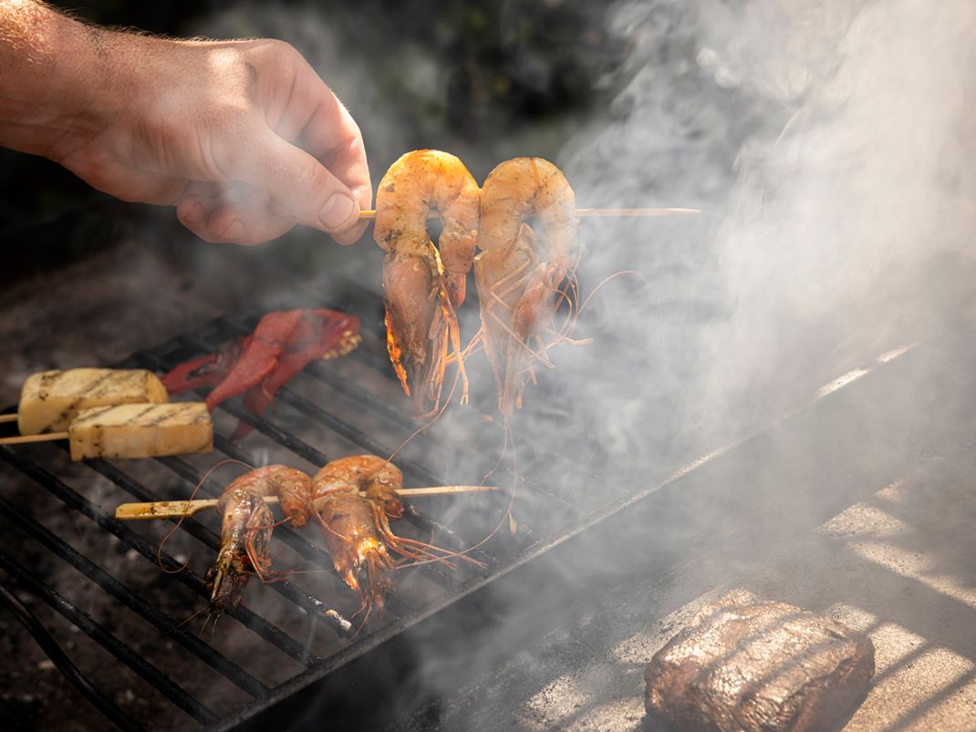 Hand holding a skewer of prawns over a smoky barbecue grill