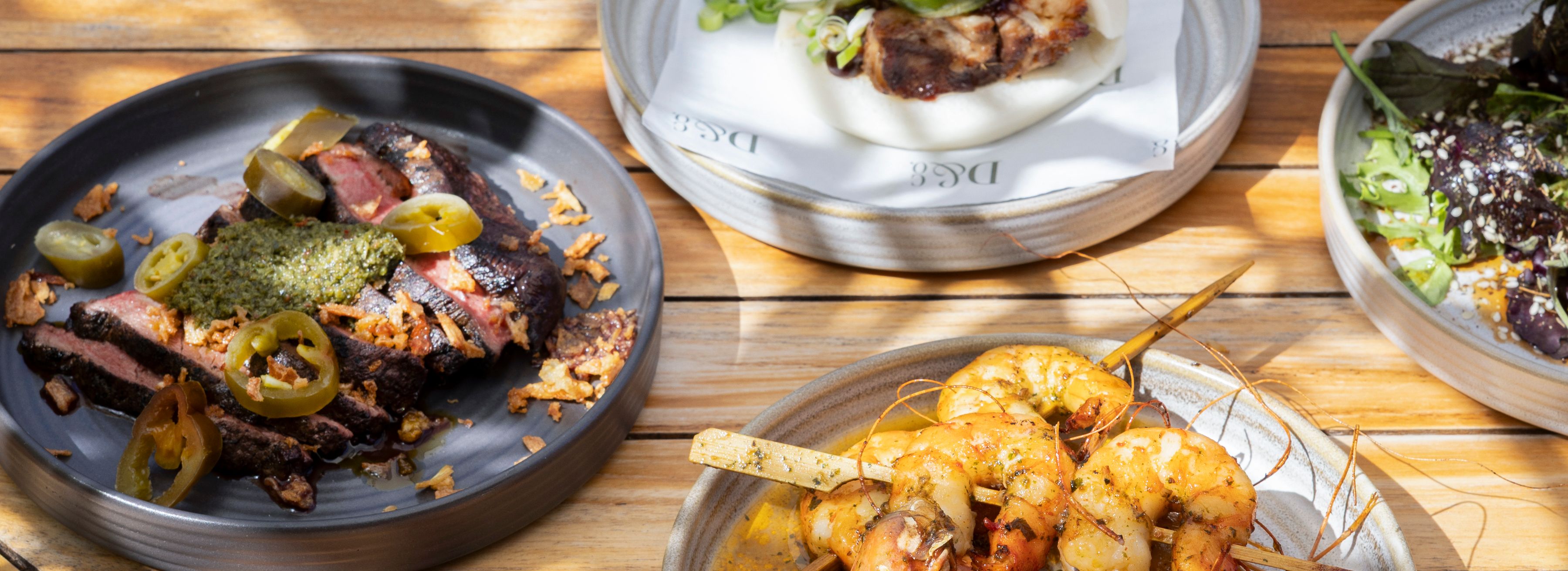 Plates of grilled prawns, sliced steak with jalapeños, a bao bun and a leafy salad on a sunlit wooden table
