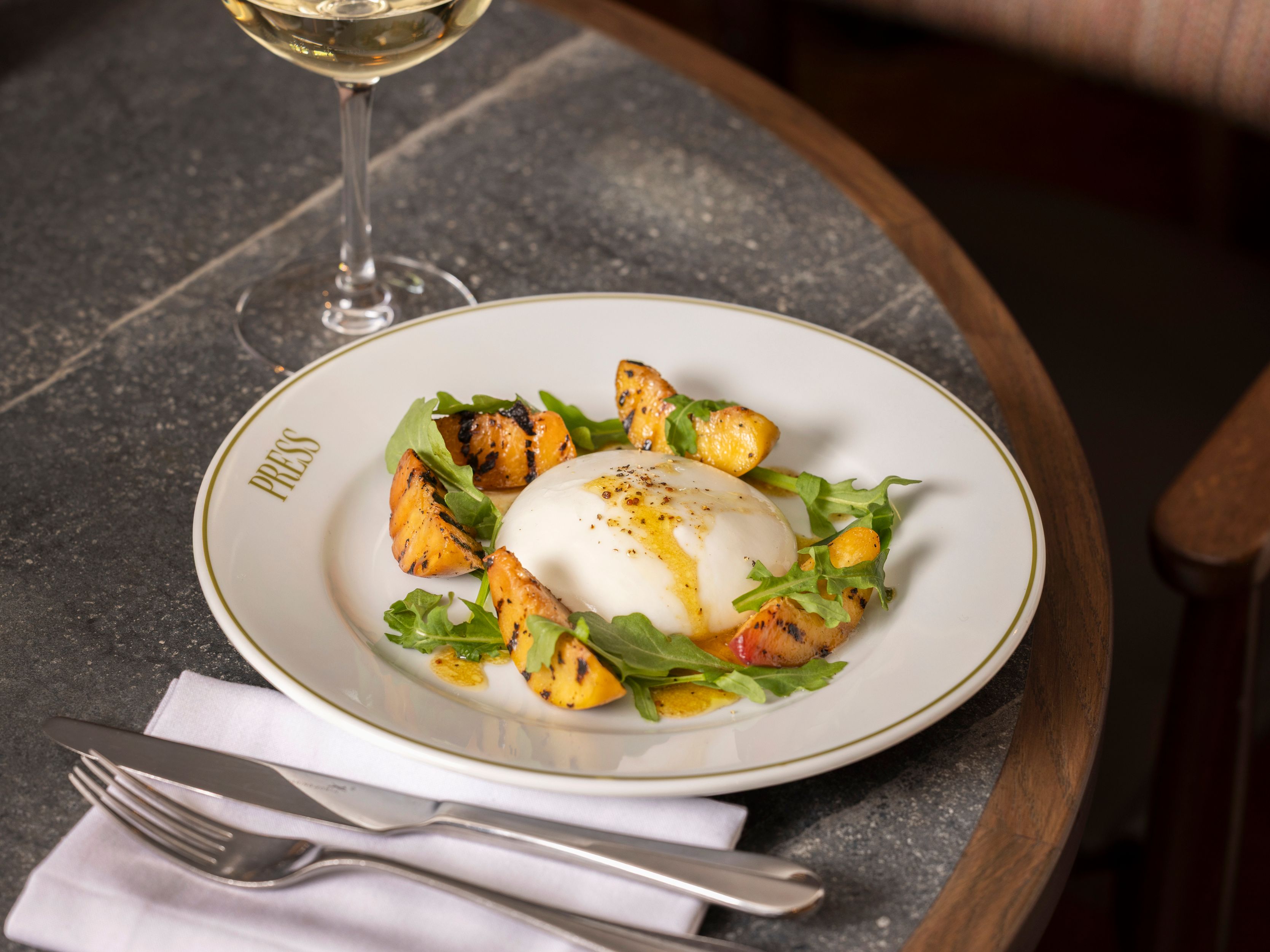 Burrata salad with grilled peaches and rocket on a white plate, with a glass of white wine on a stone table.
