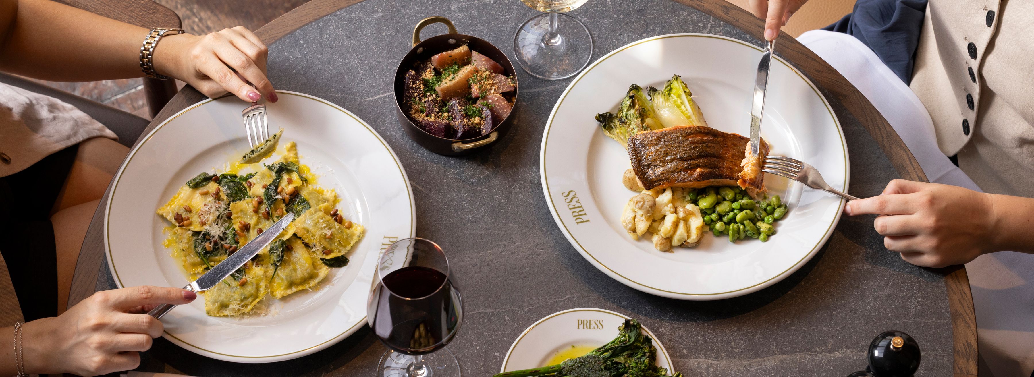 Overhead view of two people dining at a restaurant table with salmon, ravioli, vegetables and glasses of wine