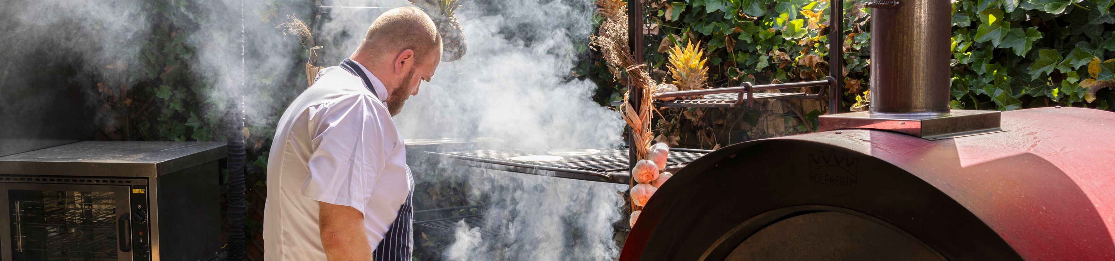 Chef tending a smoky outdoor barbecue grill beneath string lights in a leafy courtyard