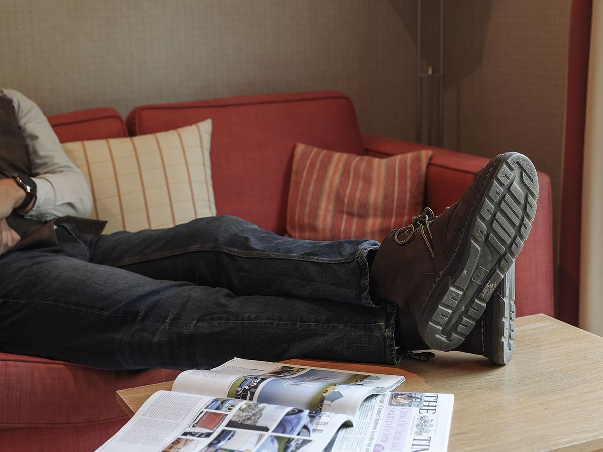Person reclining on a red sofa with boots on a wooden coffee table beside an open magazine