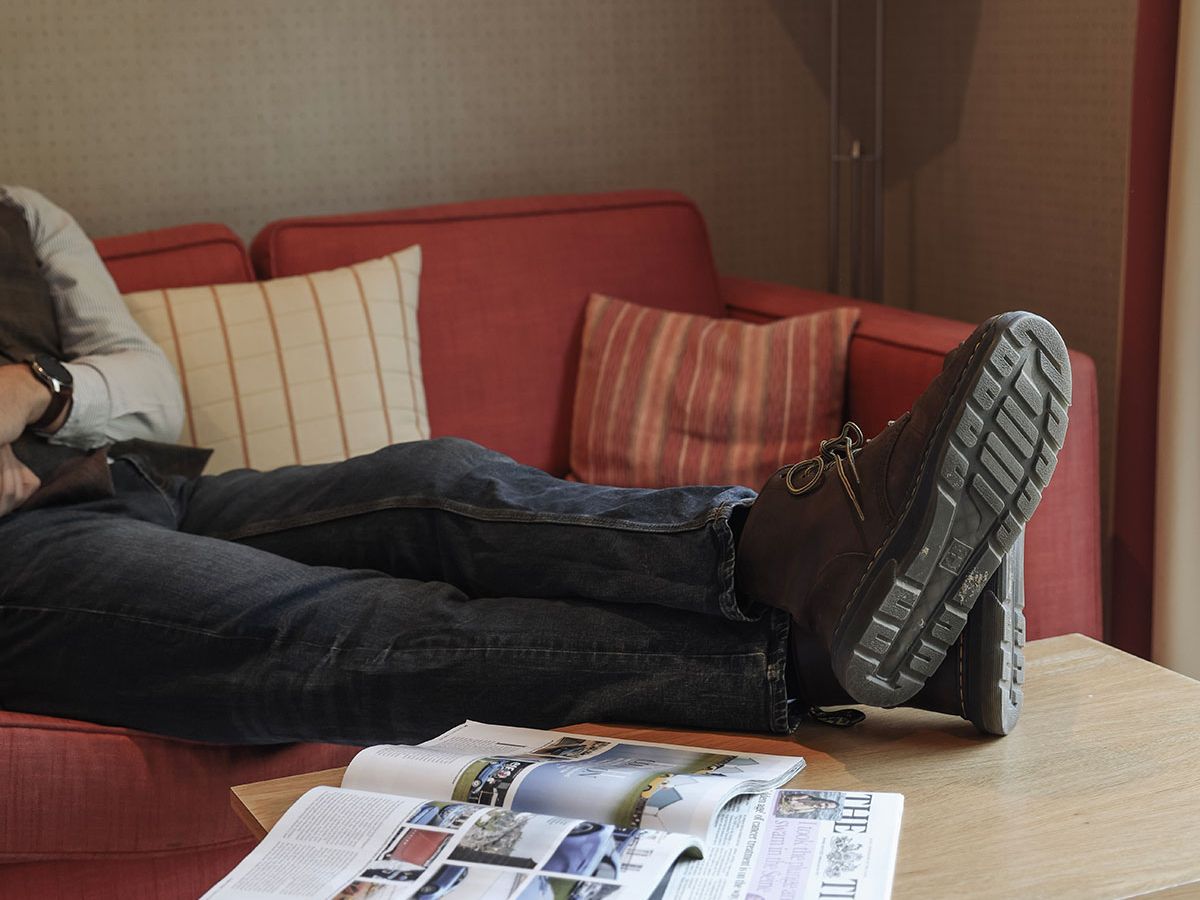 Person reclining on a red sofa with boots on a wooden coffee table beside an open magazine