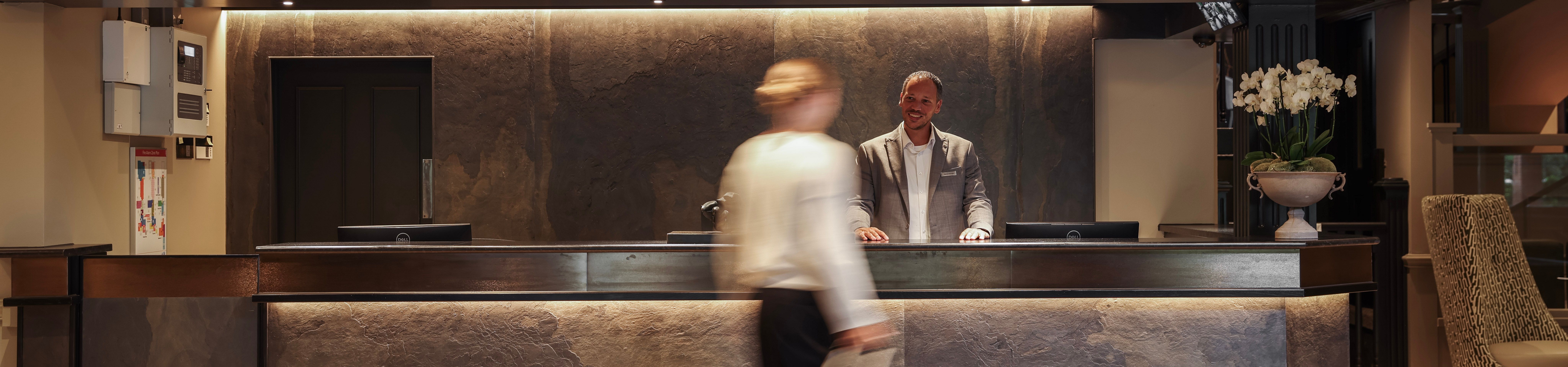 Hotel reception desk with staff member and a guest walking past with luggage