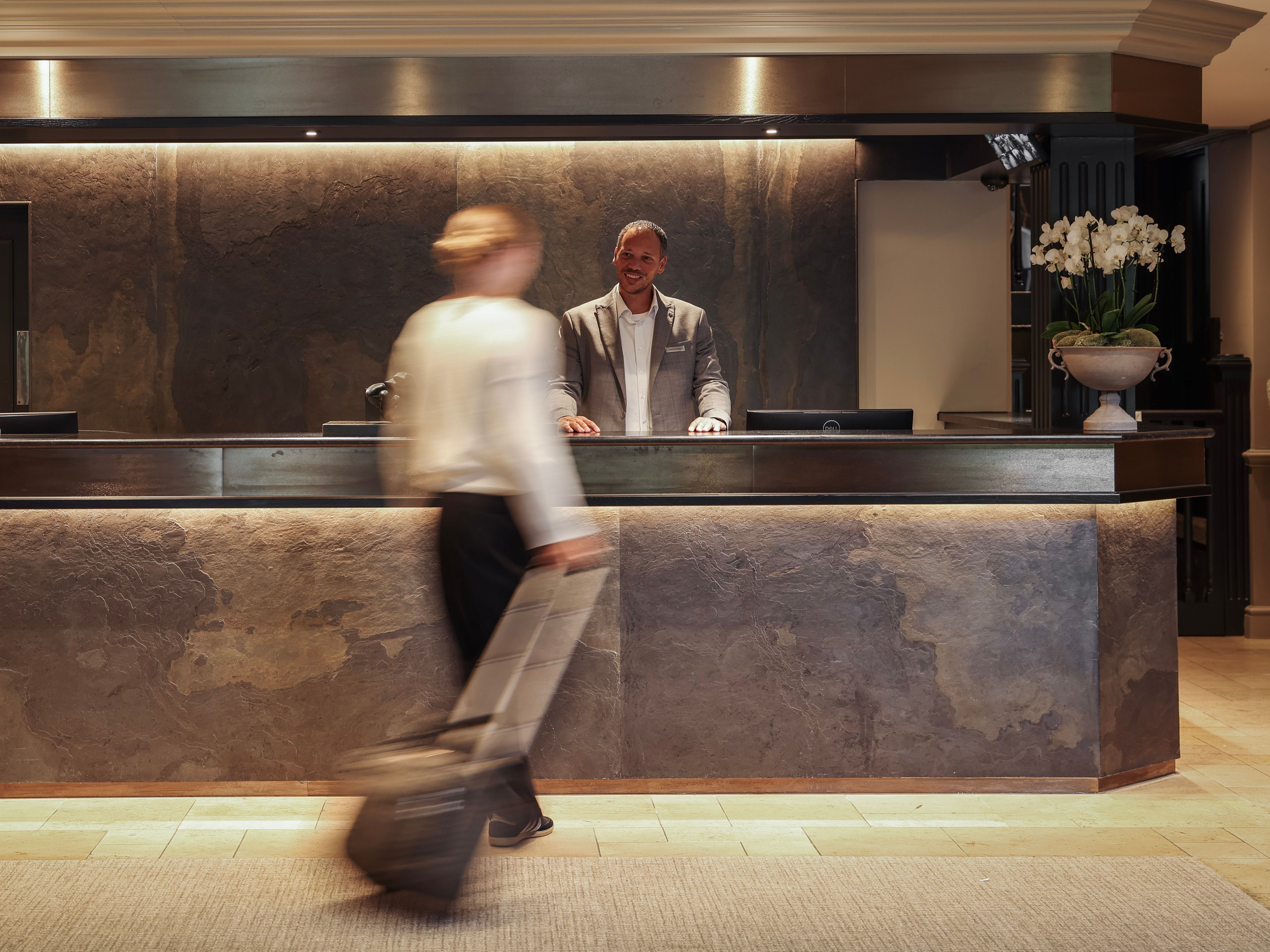 Hotel reception desk with staff member and a guest walking past with luggage