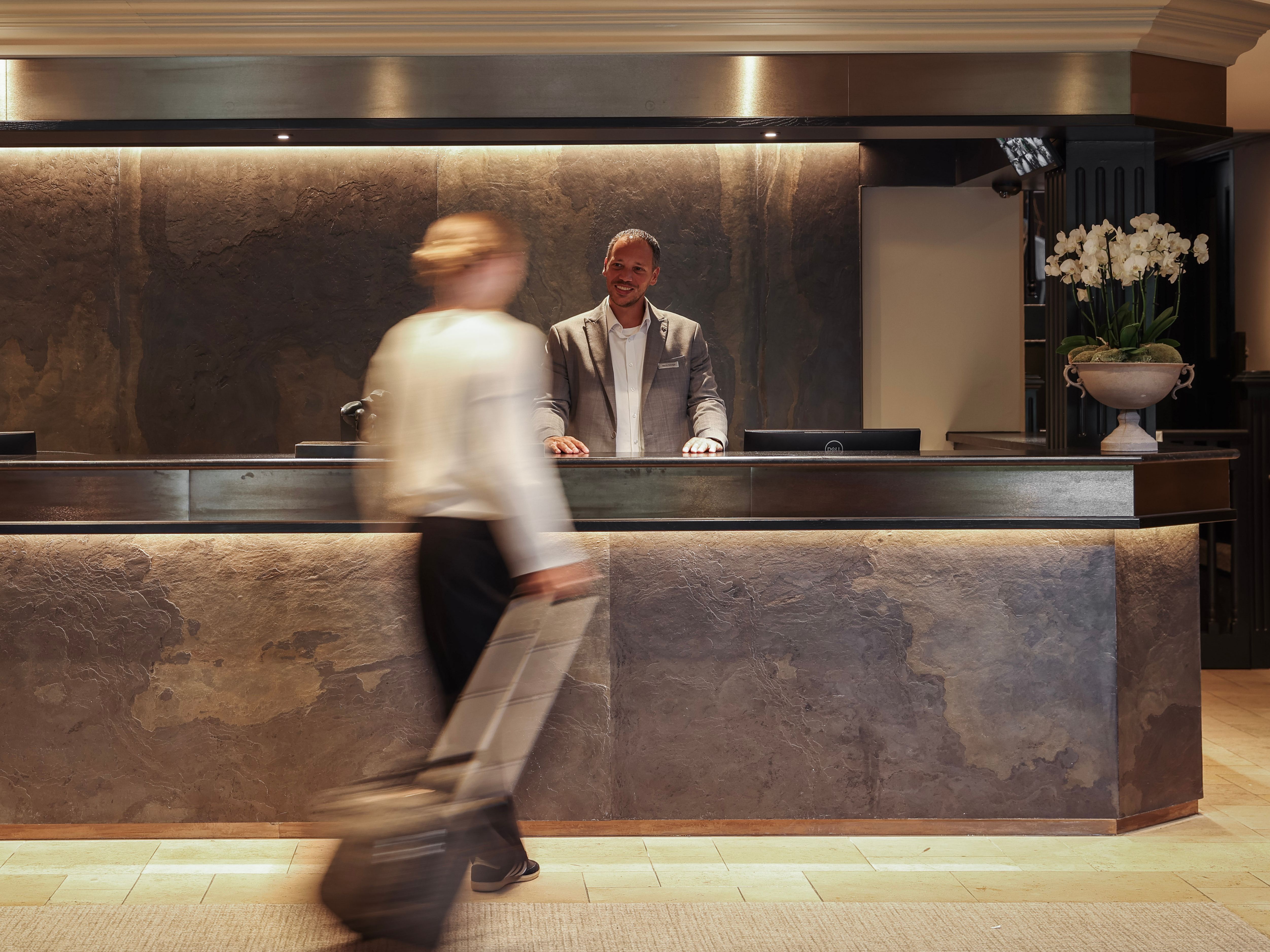 Hotel reception desk with staff member and a guest walking past with luggage