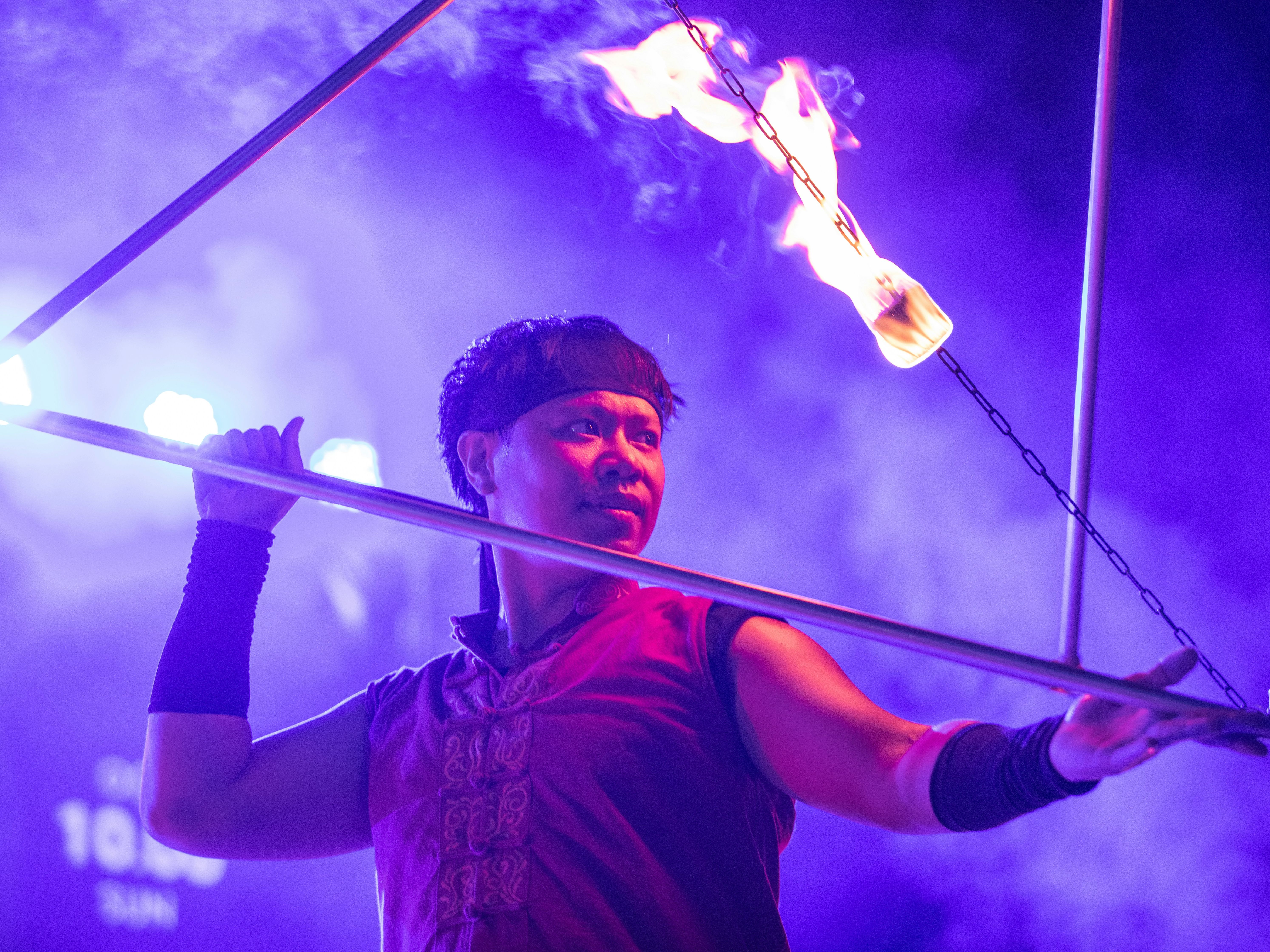 Fire performer on stage holding metal bars near a flaming torch under purple lighting