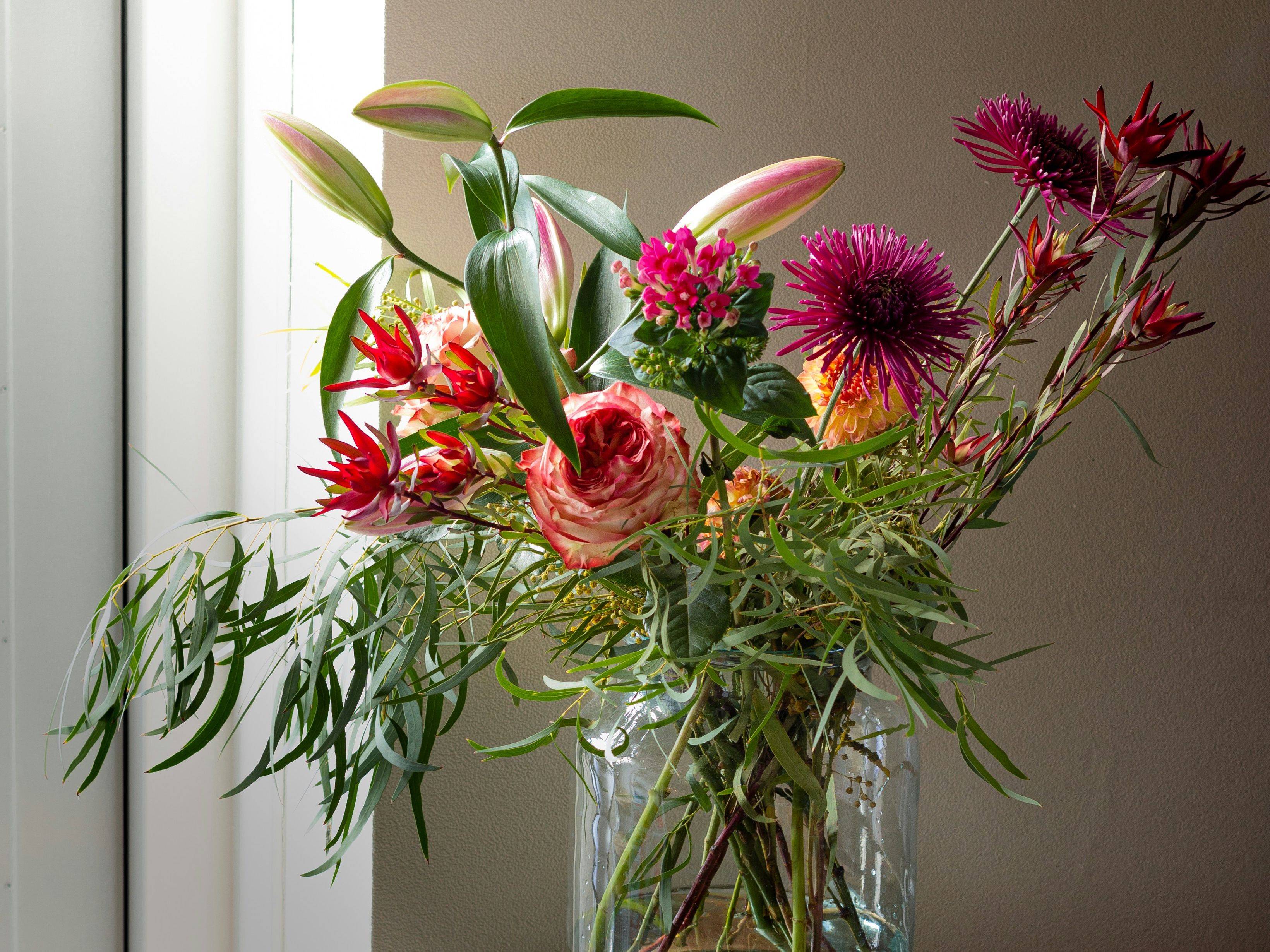 Colourful bouquet of lilies, roses and purple flowers in a glass vase on a table by a window
