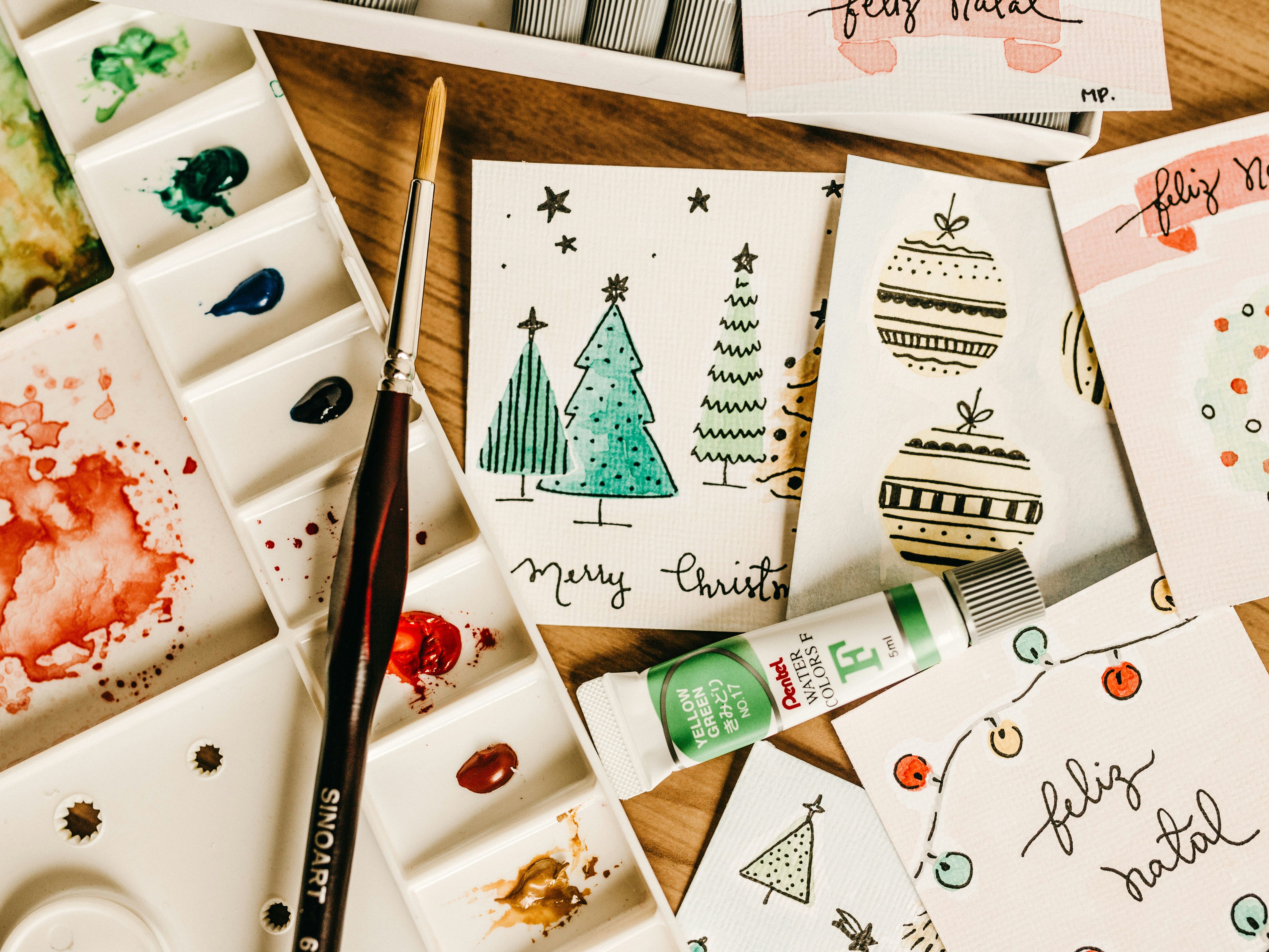 Watercolour palette and paintbrush beside handmade Christmas cards with illustrated trees and baubles on a wooden table