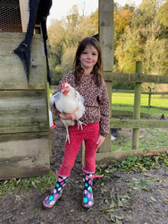 A young girl in a leopard-print sweater, red leggings, and colorful polka-dot rain boots stands in a muddy farmyard, smiling while holding a white chicken.