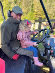 A smiling man in a green jacket and cap sits beside a young girl in pink boots and a puffy jacket as she grips the steering wheel of a utility vehicle on a farm.