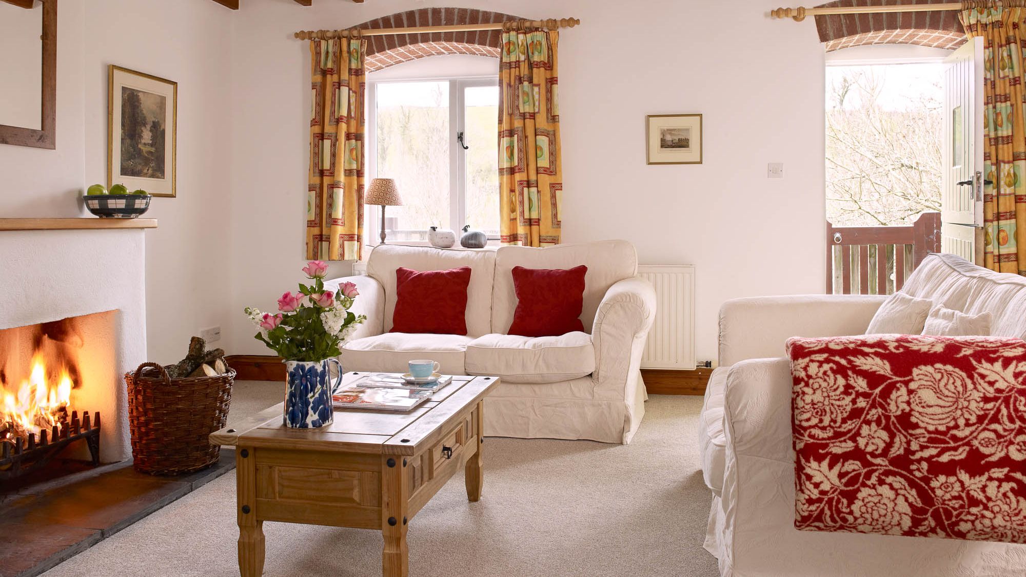 A cozy living room with white sofas, red accent pillows, and a roaring fireplace. A wooden coffee table with flowers sits in the centre, and an open door leads outside.