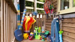 An outdoor storage area with sports and beach equipment, including tennis rackets, golf clubs, buckets, shovels, and inflatable toys, neatly arranged against a wooden wall.