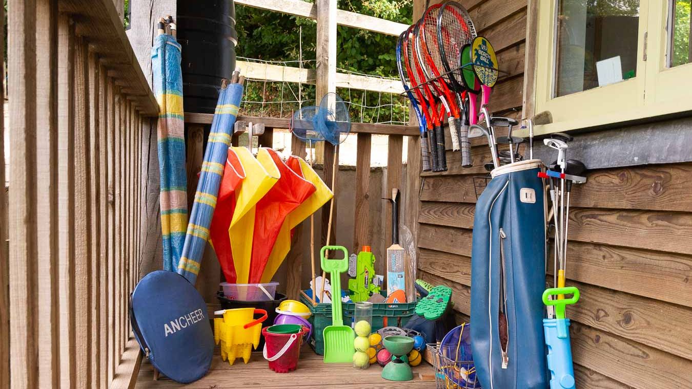 An outdoor storage area with sports and beach equipment, including tennis rackets, golf clubs, buckets, shovels, and inflatable toys, neatly arranged against a wooden wall.