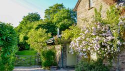 A charming stone cottage with a green door, surrounded by lush greenery and blooming pink roses, set against a backdrop of trees and a wooden gate leading to a garden.