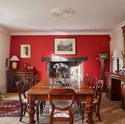 A luxurious dining room with a rich red accent wall, a long wooden table set with glassware, antique furniture, a grand mirror, and a rustic fireplace.