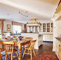 A bright, country-style kitchen with a large wooden dining table set for breakfast, cream cabinetry, an Aga stove, and rustic accents, bathed in natural light.