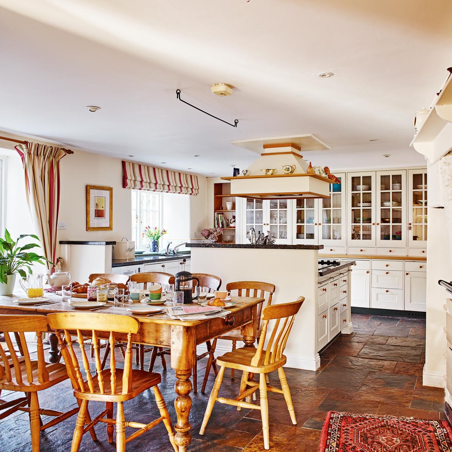 A bright, country-style kitchen with a large wooden dining table set for breakfast, cream cabinetry, an Aga stove, and rustic accents, bathed in natural light.